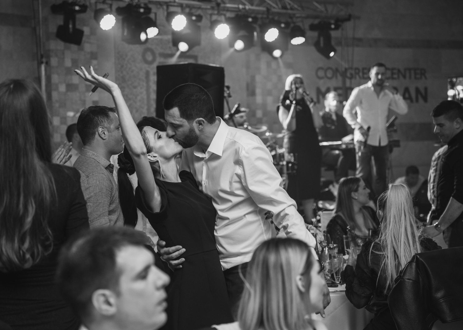 Couple sharing romantic kiss on crowded wedding dance floor with live band performing on stage.