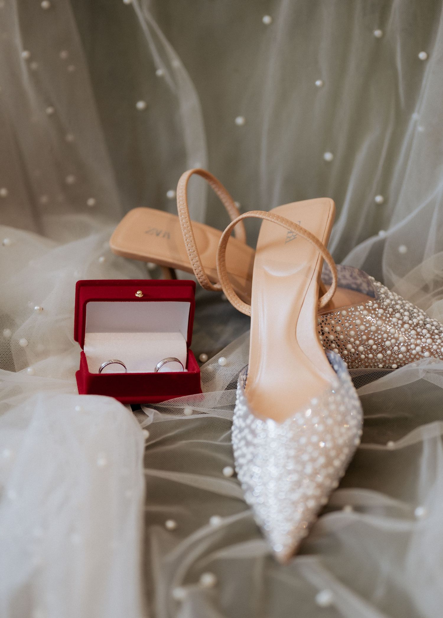 Close-up of pearl-embellished bridal heels beside a red ring box with wedding bands on veil.