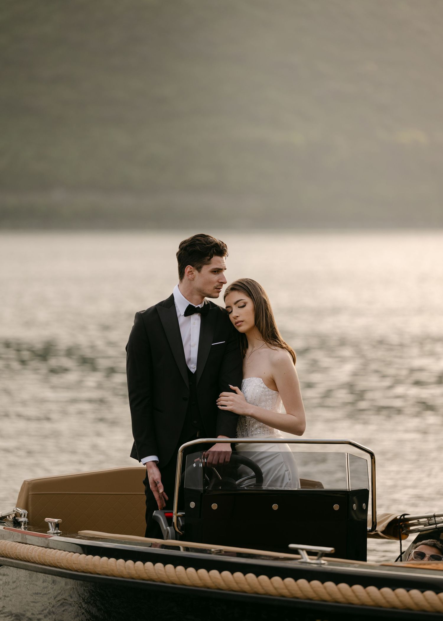 Elegant wedding couple posing on a classic wooden boat at golden hour with misty mountains behind them.
