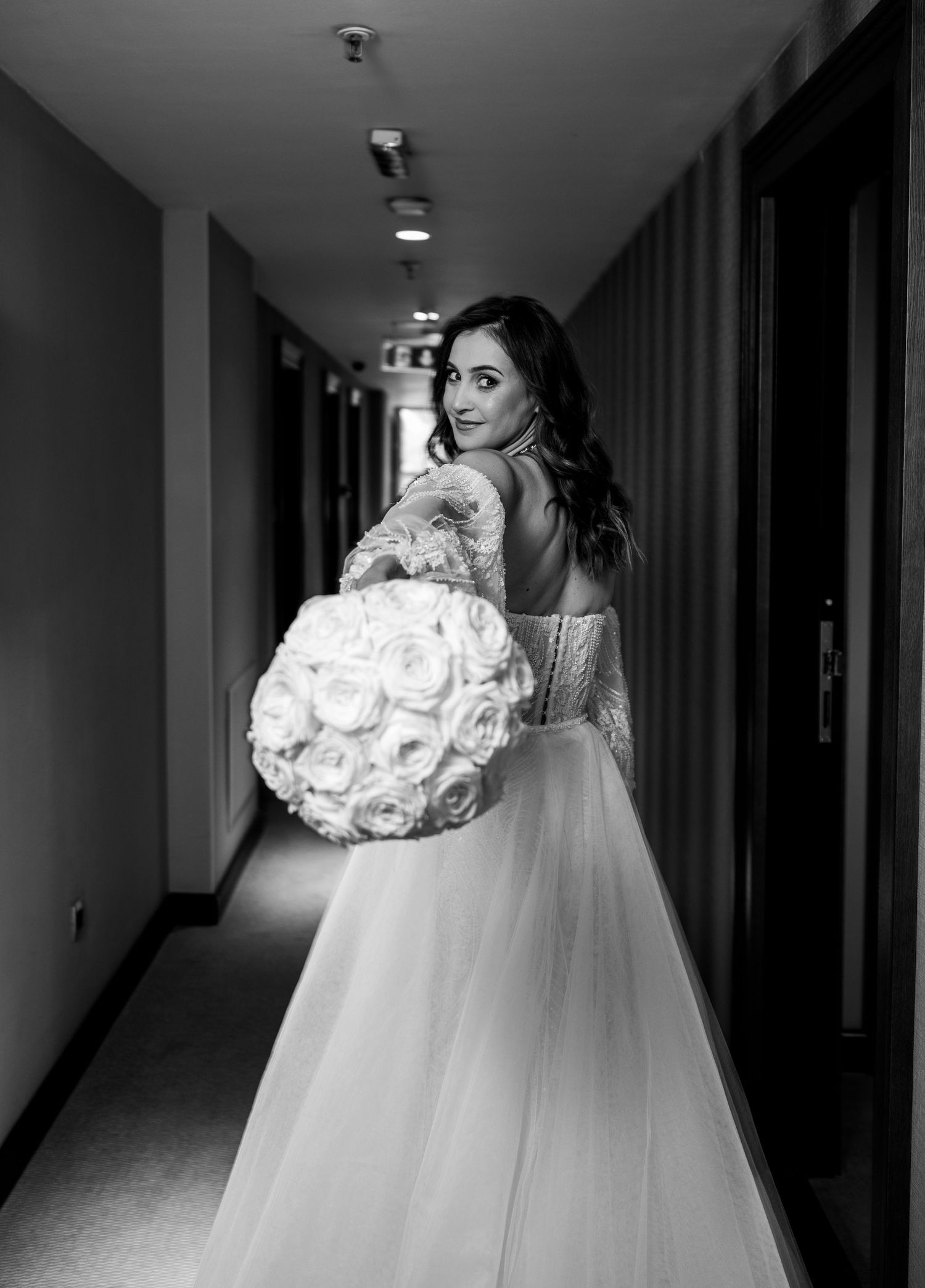 Black and white photo of bride in backless wedding gown holding round rose bouquet in hotel hallway.