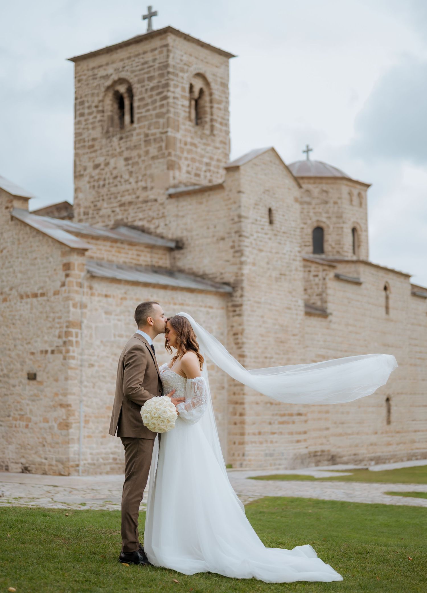 Bride and groom kissing outside a stone church, brides veil flowing in wind, holding white bouquet.