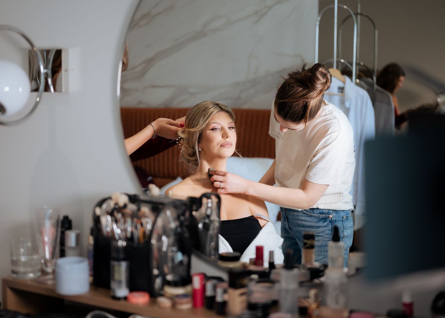 Bride getting makeup applied at vanity mirror surrounded by cosmetics before wedding.