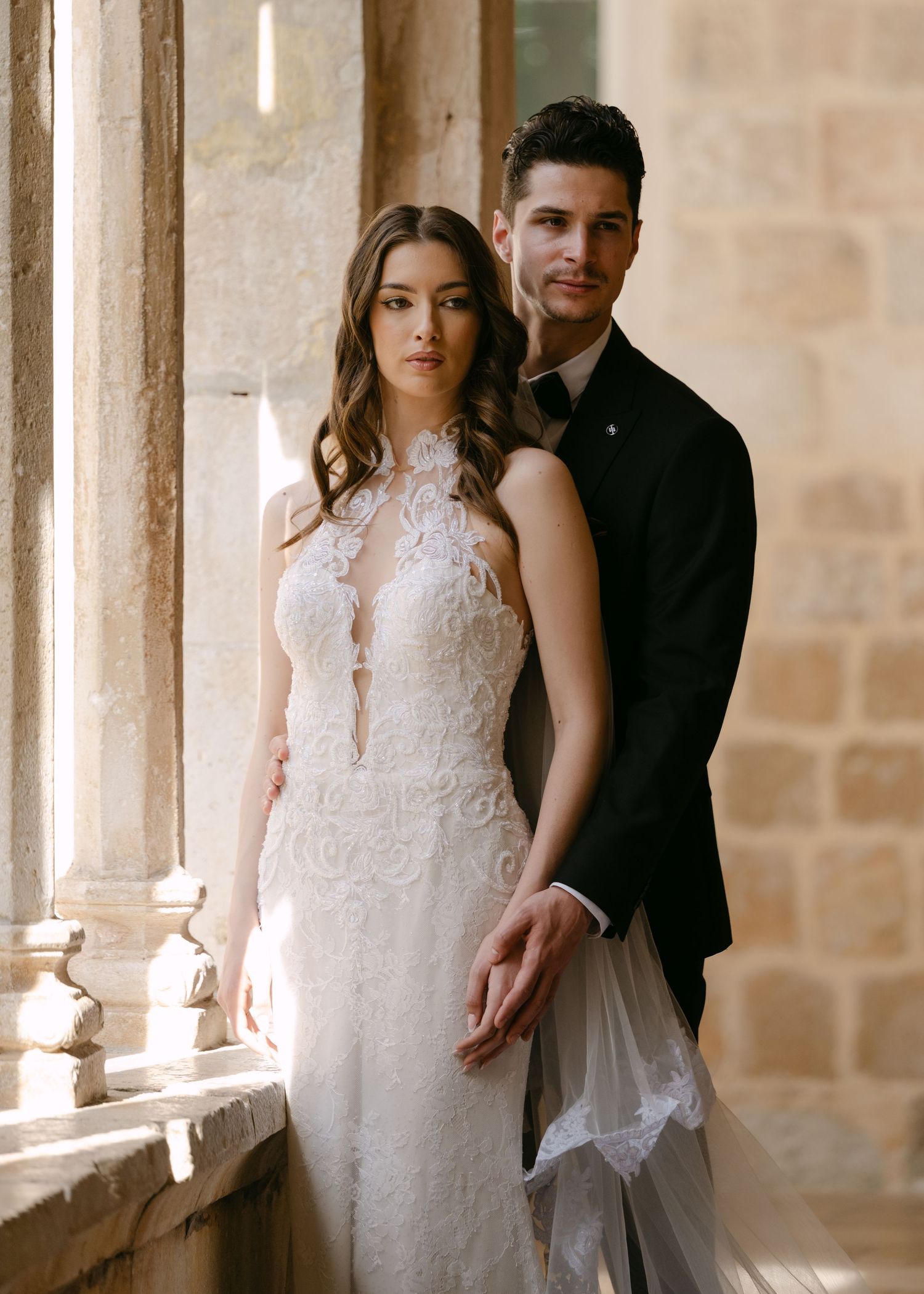 Elegant bride and groom in formal wedding attire standing together by sunlit stone columns.