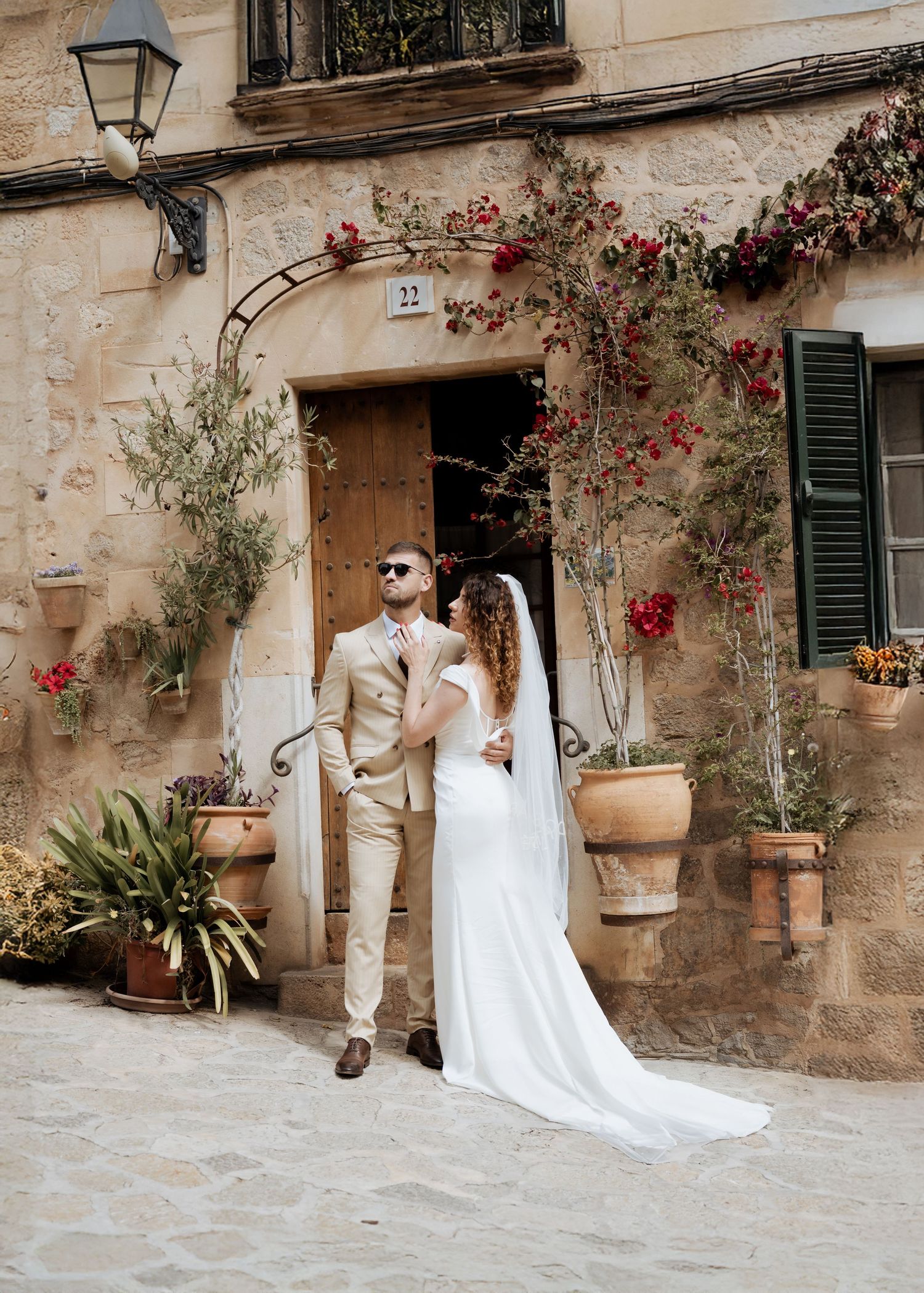 Wedding couple stands before rustic stone building adorned with red flowers in cobblestone courtyard.