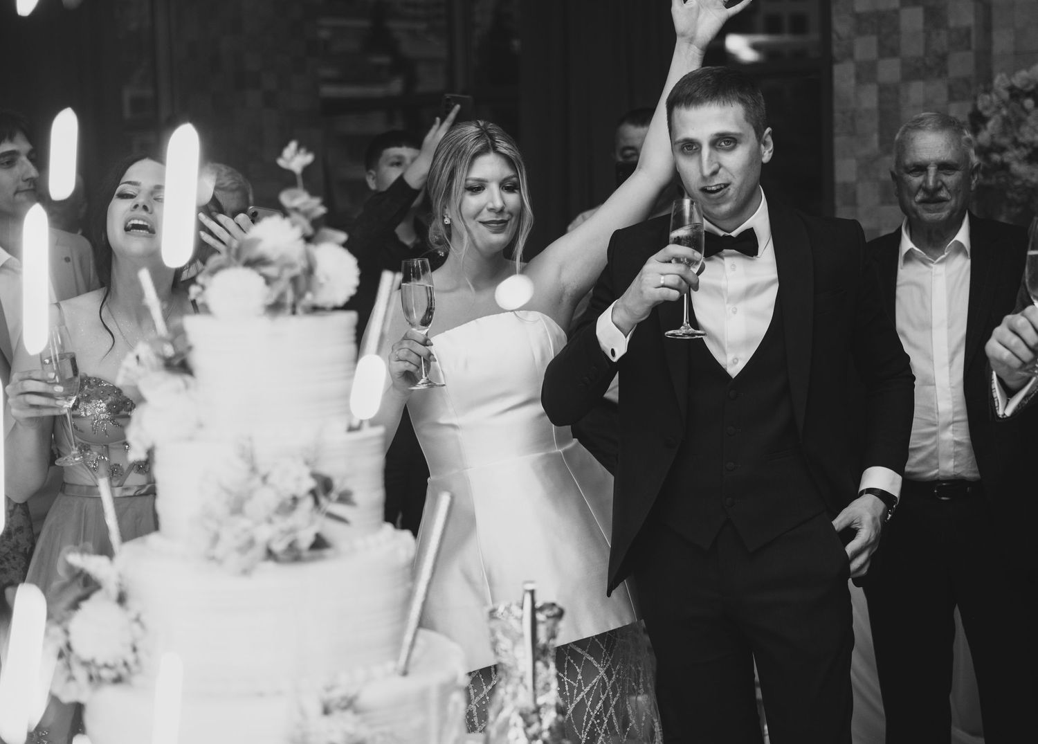 Black and white photo of bride and groom at wedding reception near tiered cake with sparklers and guests.