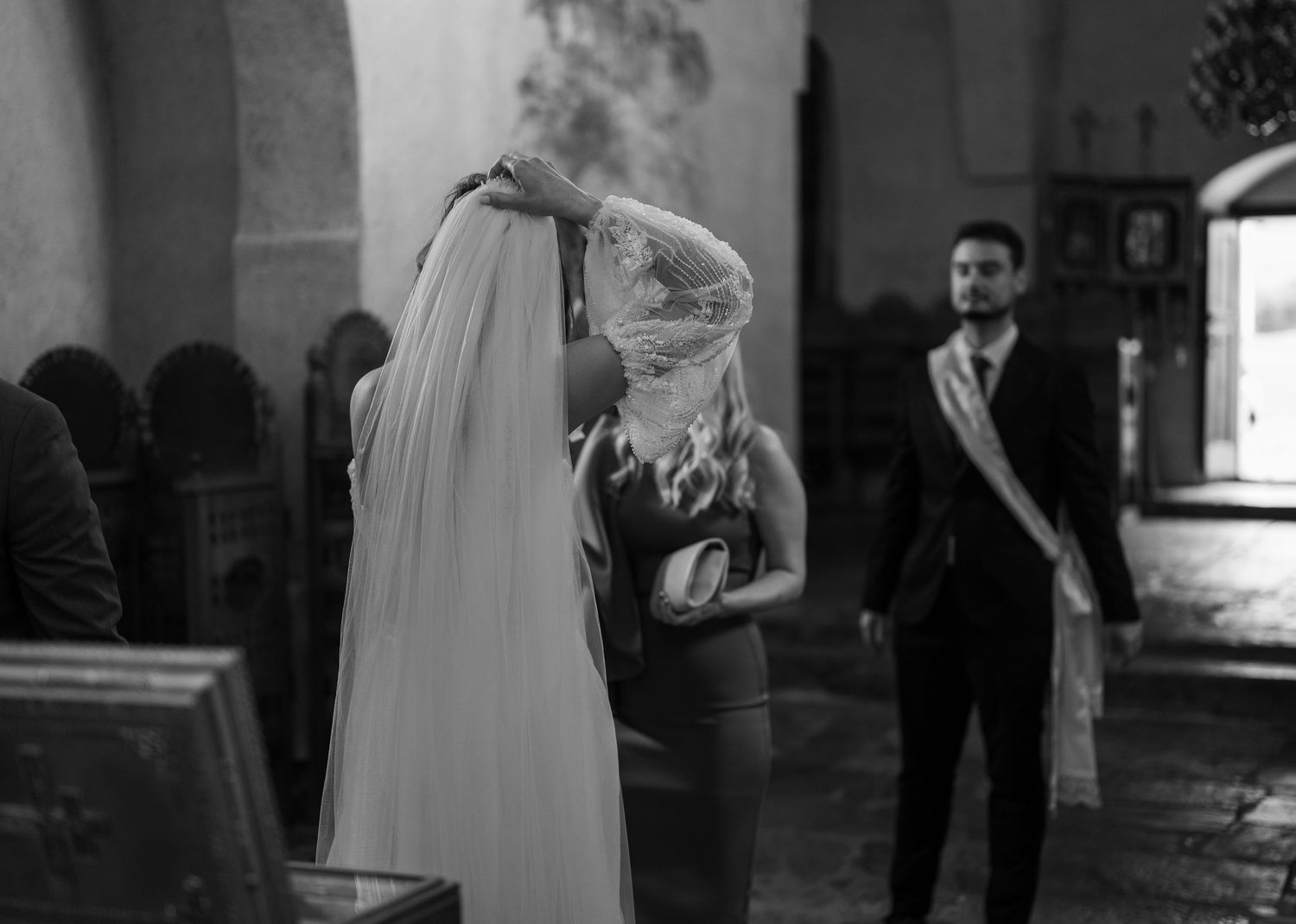 Black and white photo of bride receiving veil adjustment inside historic stone church ceremony.