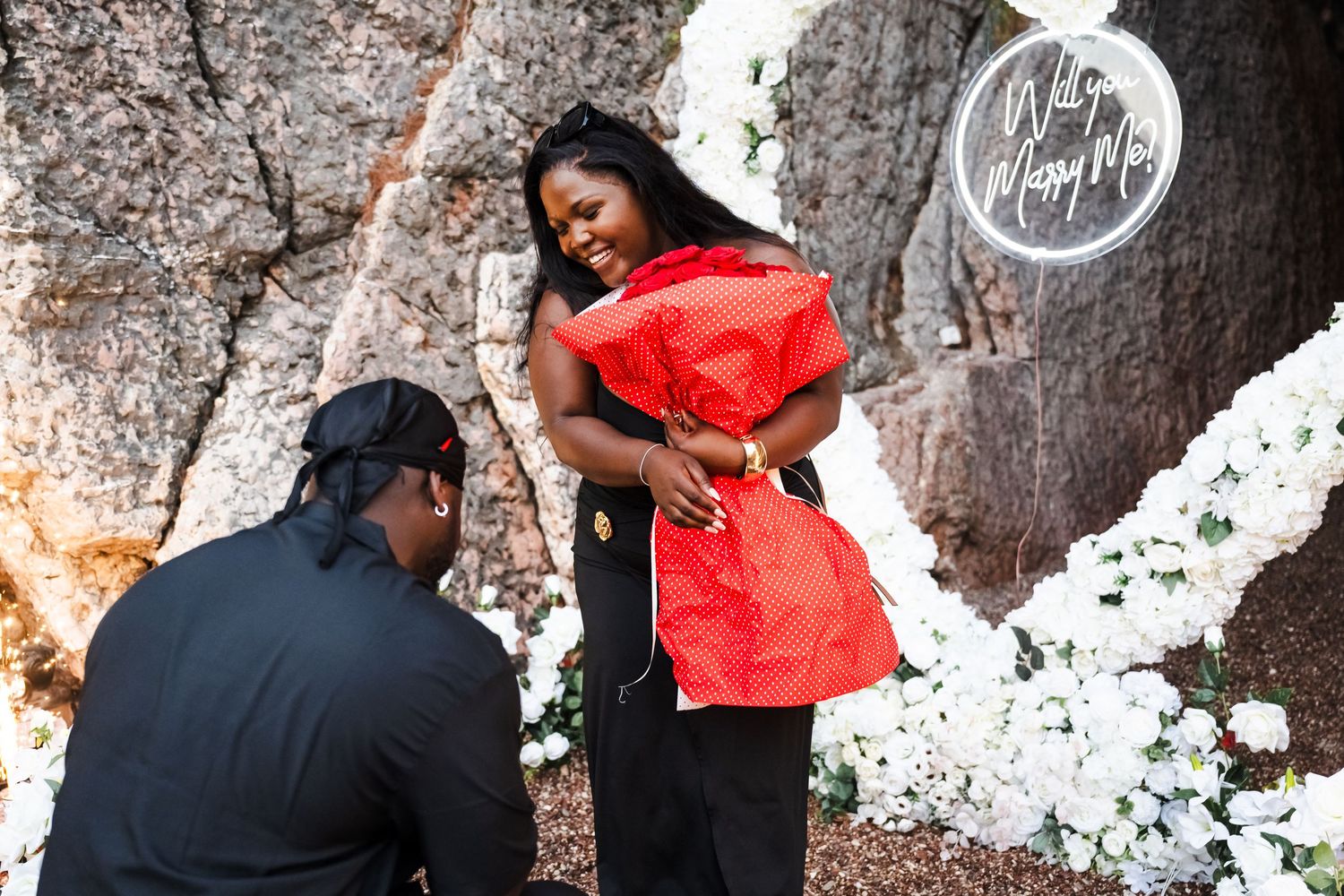 A man proposes to a woman kneeling before a Will You Marry Me neon sign and white flowers.