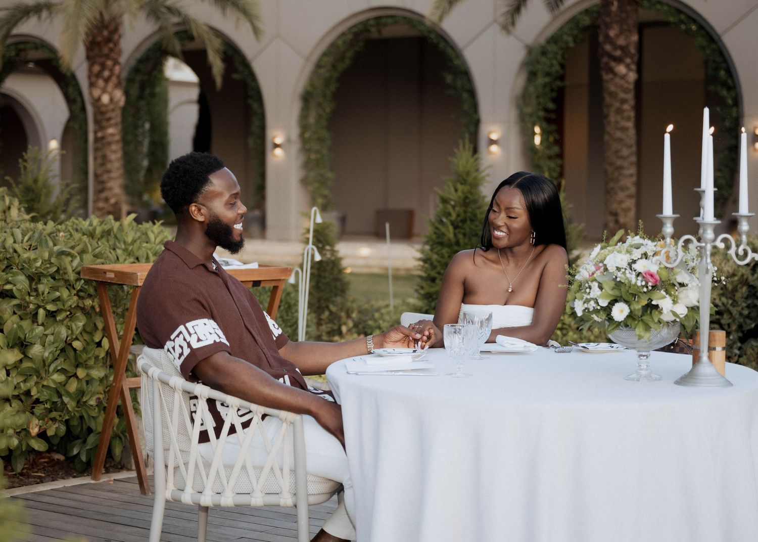 A smiling couple shares a candlelit outdoor dinner surrounded by palm trees and floral arrangements.