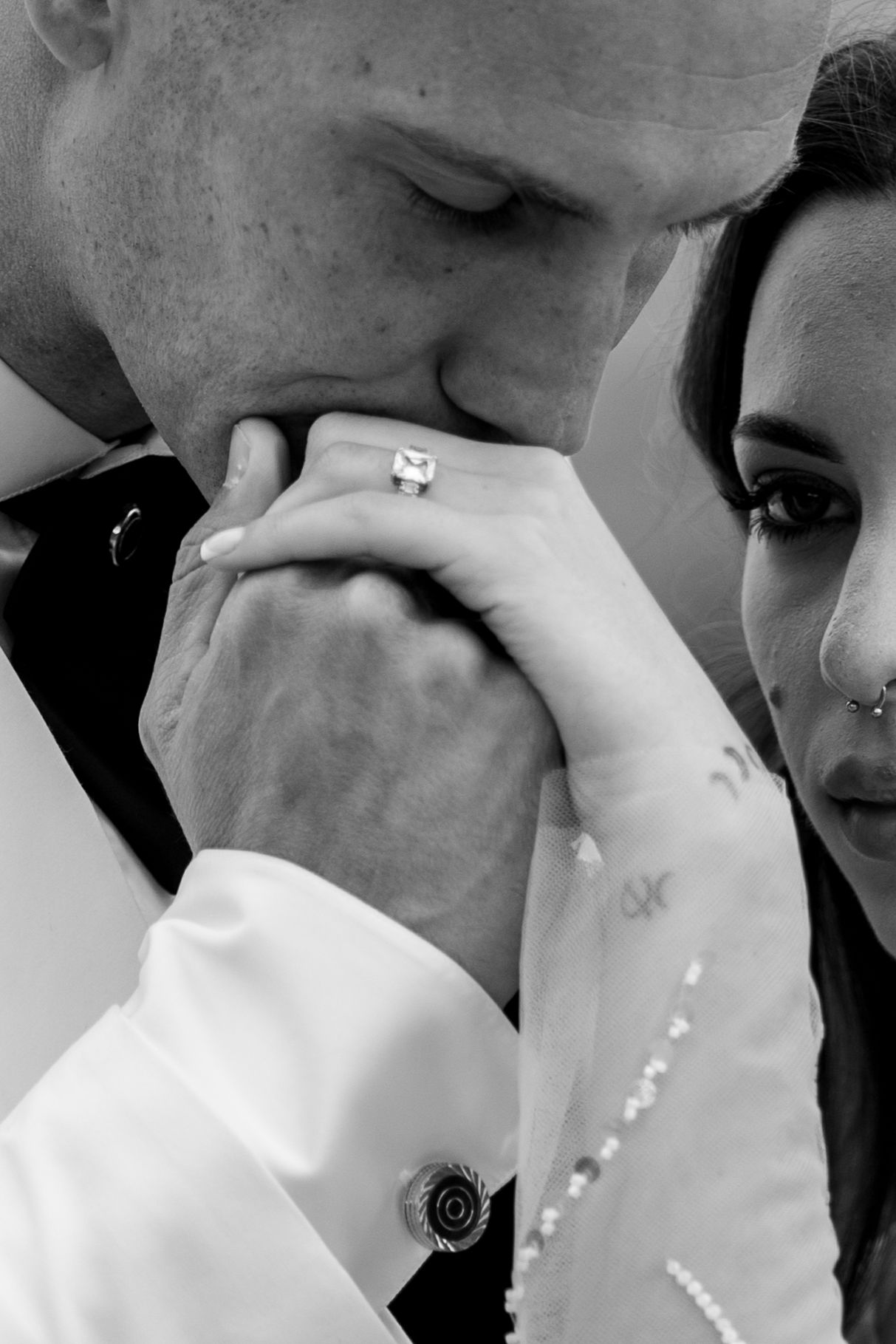 Groom kissing bride's hand adorned with diamond ring in romantic black and white wedding portrait.