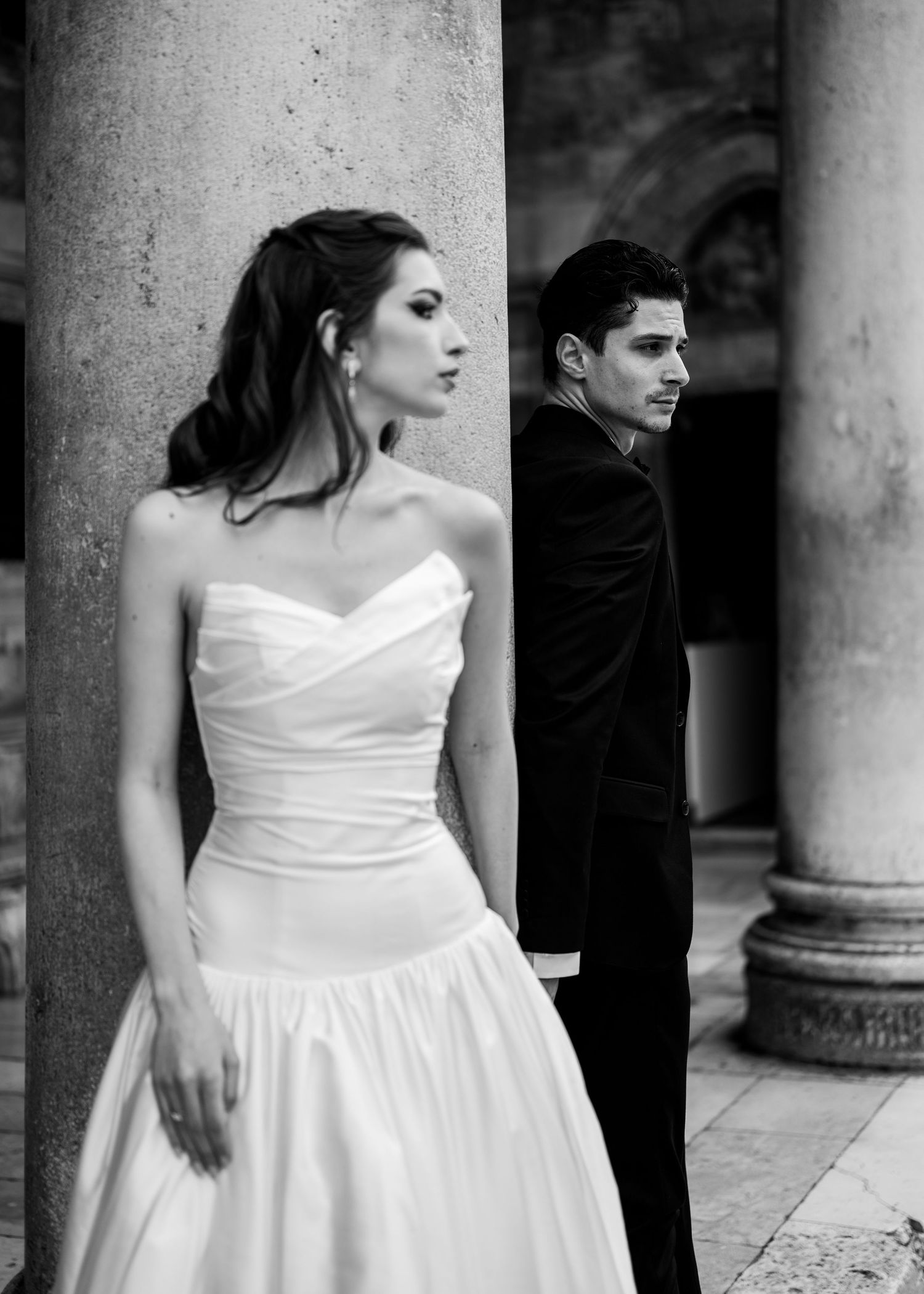 Black and white portrait of bride in strapless gown and groom leaning against stone columns looking away.