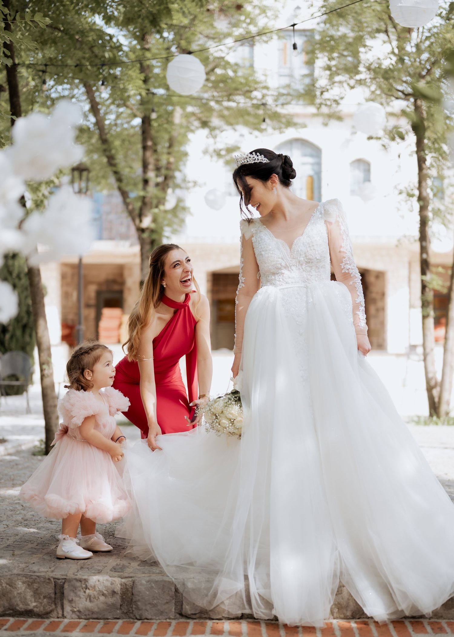 Bride in white gown with bridesmaid in red dress and flower girl in pink tutu at outdoor wedding venue.