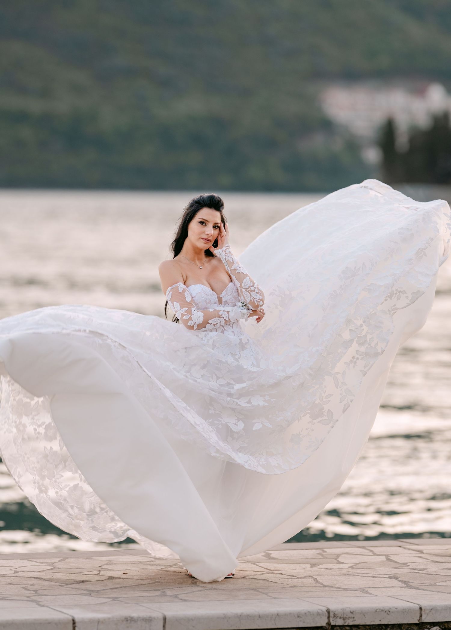 Bride in stunning white lace off-shoulder gown with flowing skirt blowing in wind by the sea.