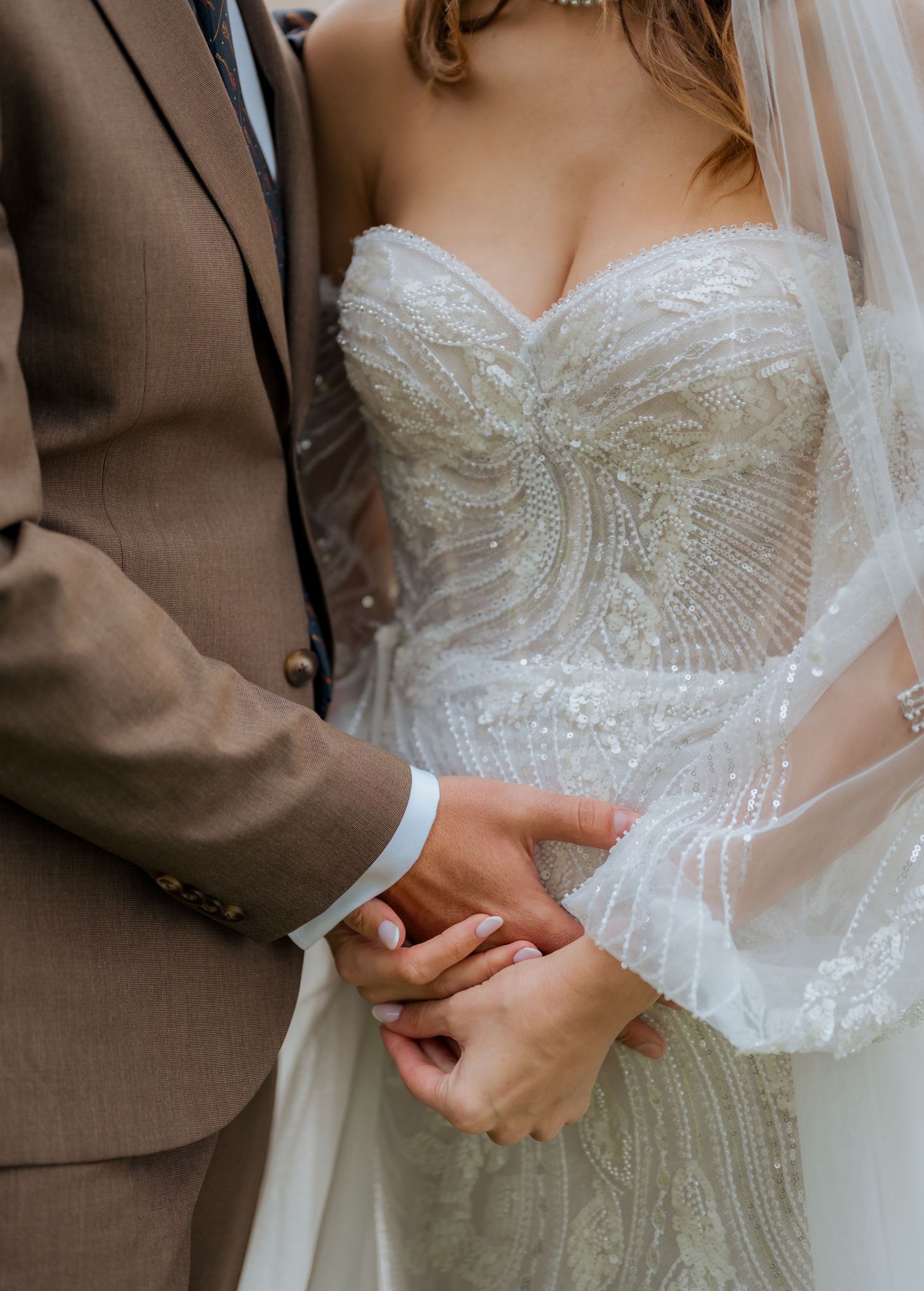 Close-up of bride and groom holding hands, bride wearing beaded strapless wedding gown with veil, groom in tan suit.