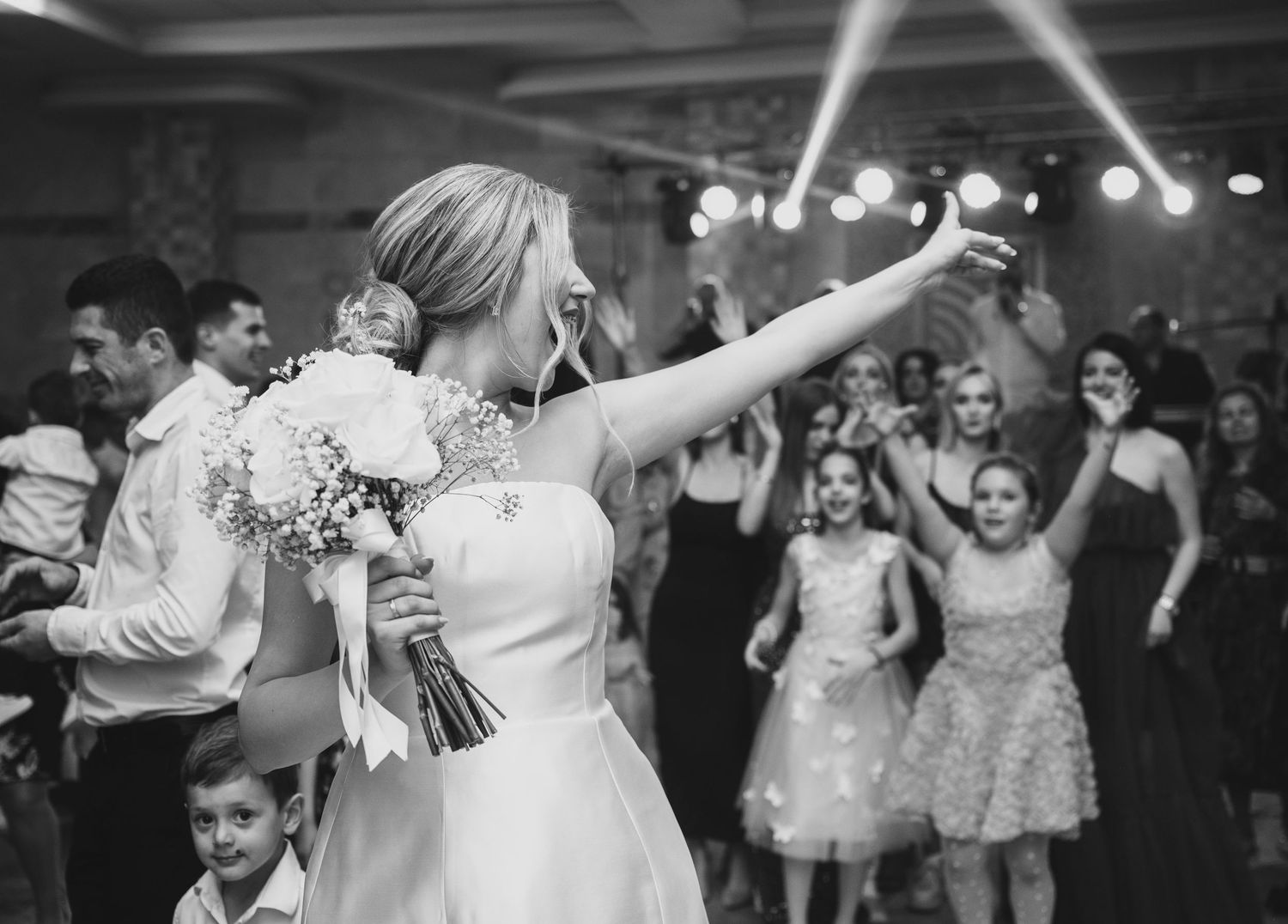 Bride in white gown tossing bouquet at wedding reception while guests celebrate on dance floor.