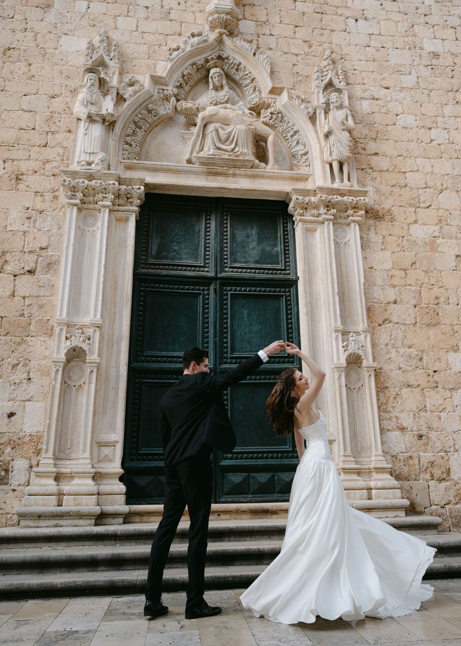Bride and groom dancing before an ornate green door adorned with elaborate stone carvings and religious sculptures.