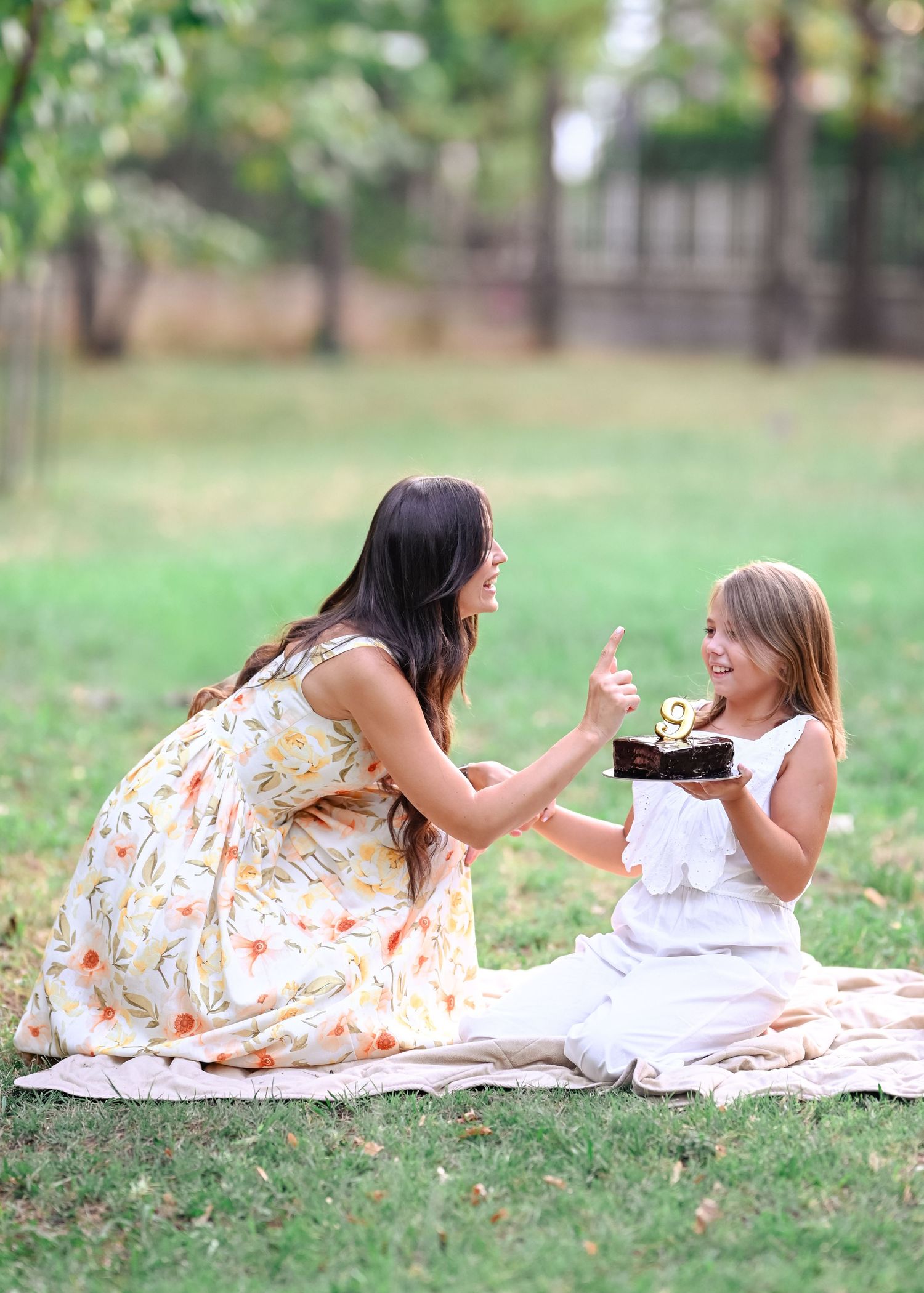 A woman in a floral dress shares a chocolate cake with a smiling young girl on a blanket in a park.