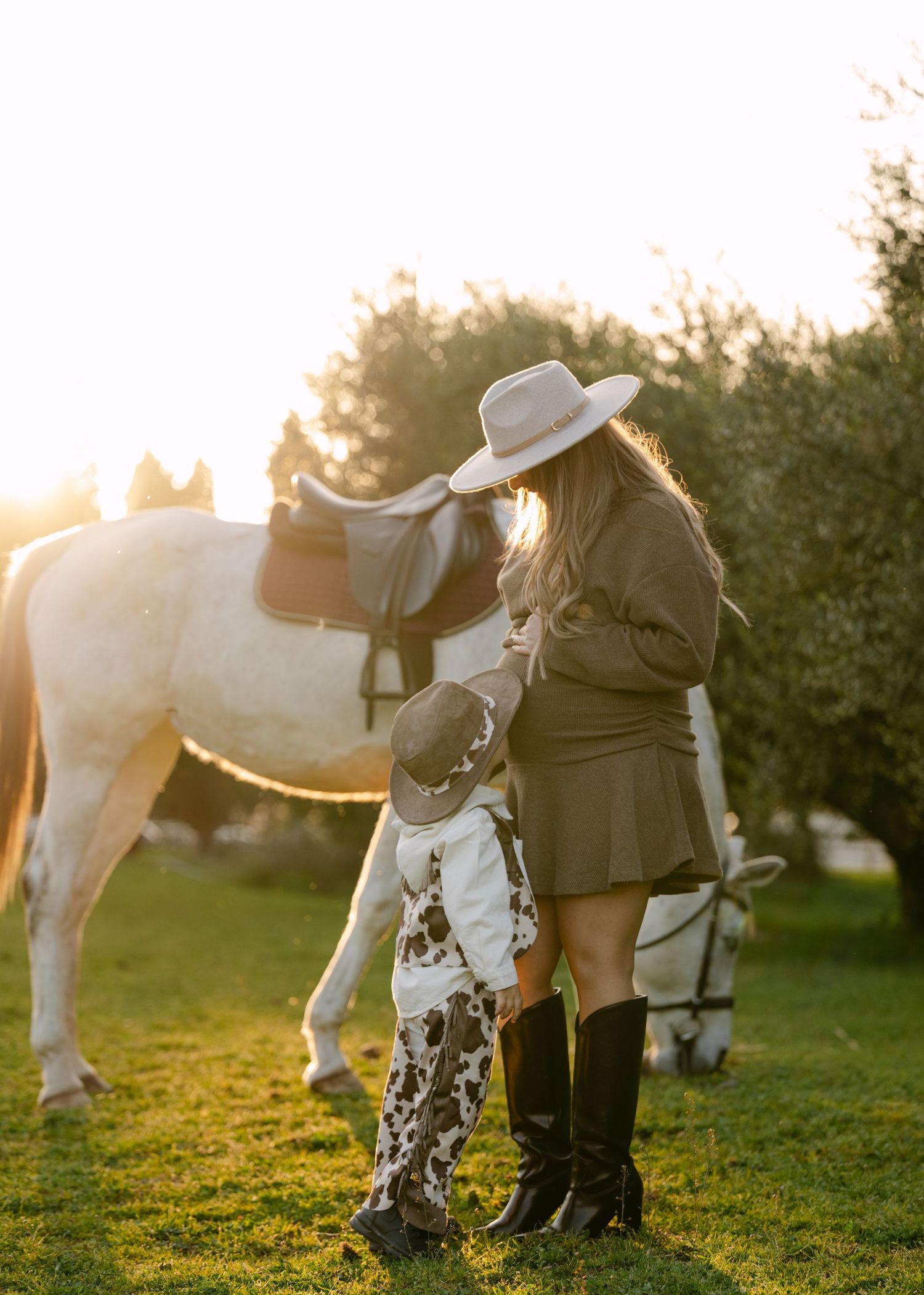 Woman and small child in cowboy outfits standing beside saddled white horse at sunset.