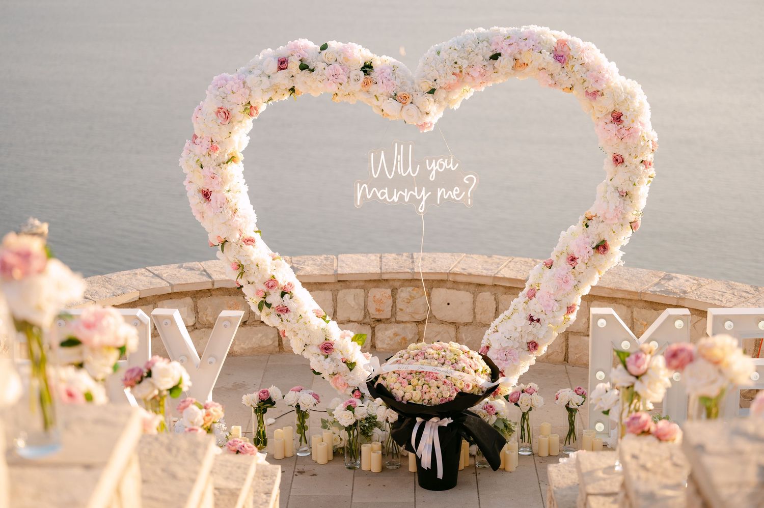 Elegant outdoor proposal setup with a floral heart arch and Will You Marry Me sign by the sea.