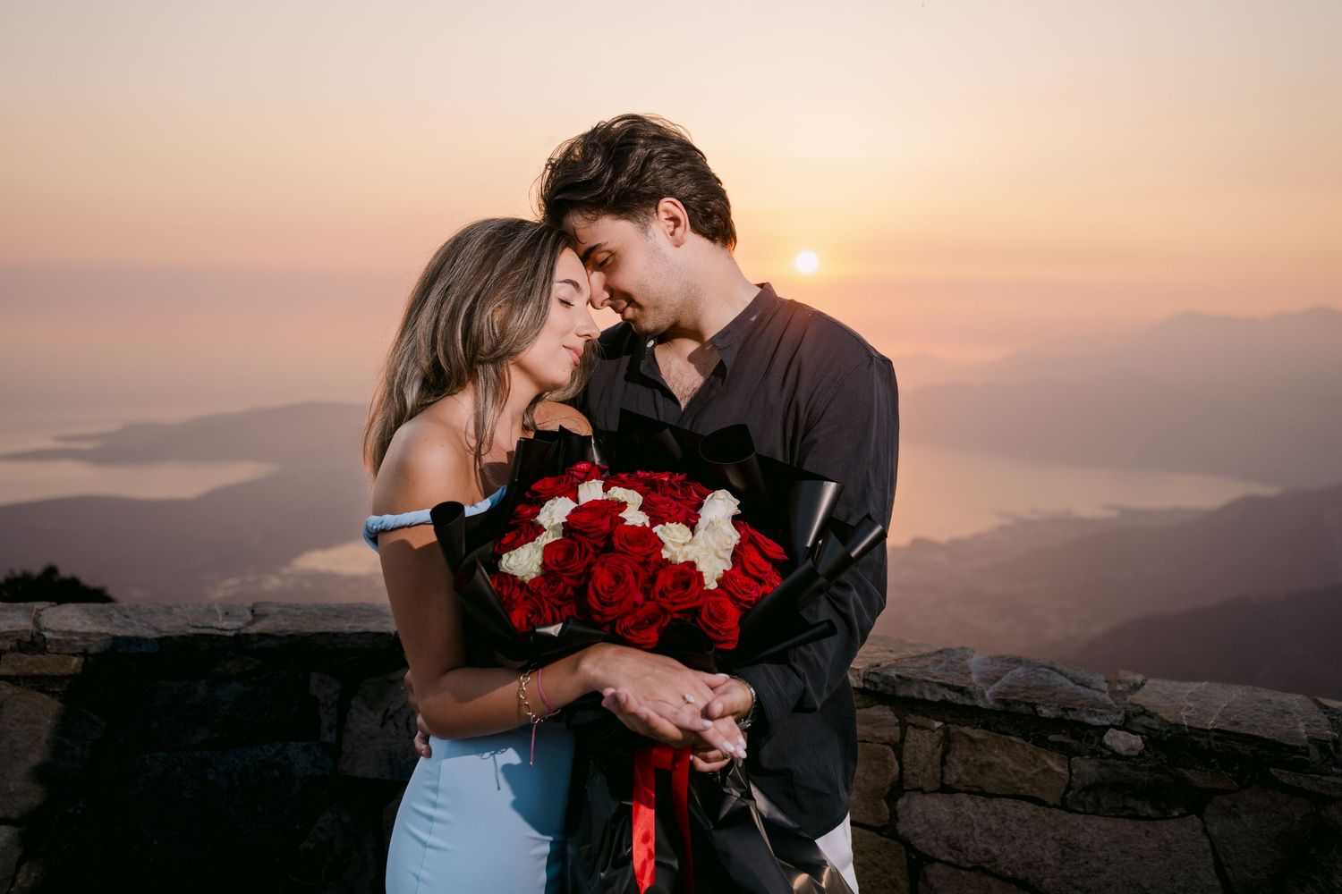 Romantic couple holding red and white roses bouquet at sunset with mountain lake views behind them.