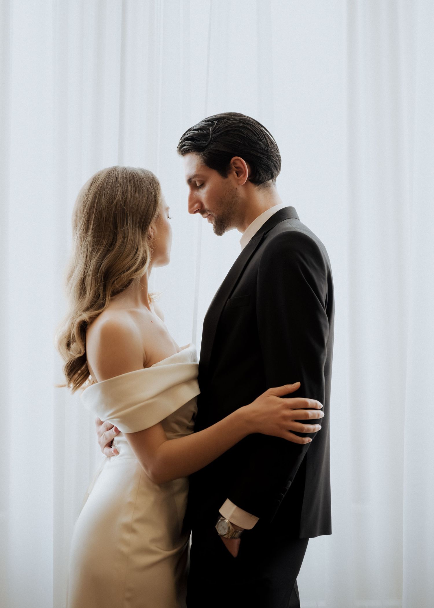 Romantic couple in wedding attire embracing closely before sheer white curtains in soft natural light.