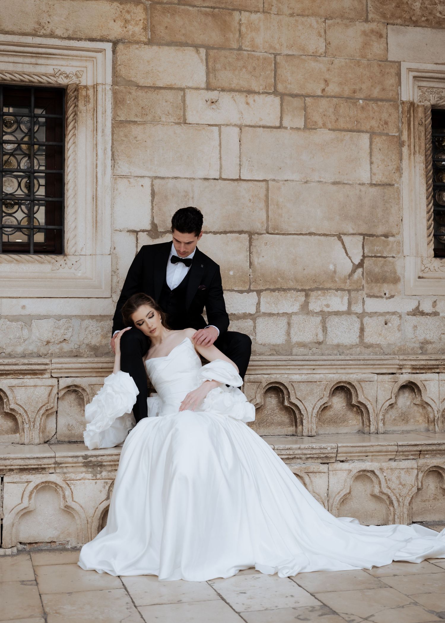 Groom in black tuxedo tenderly holding bride in a voluminous white gown on ornate stone steps.