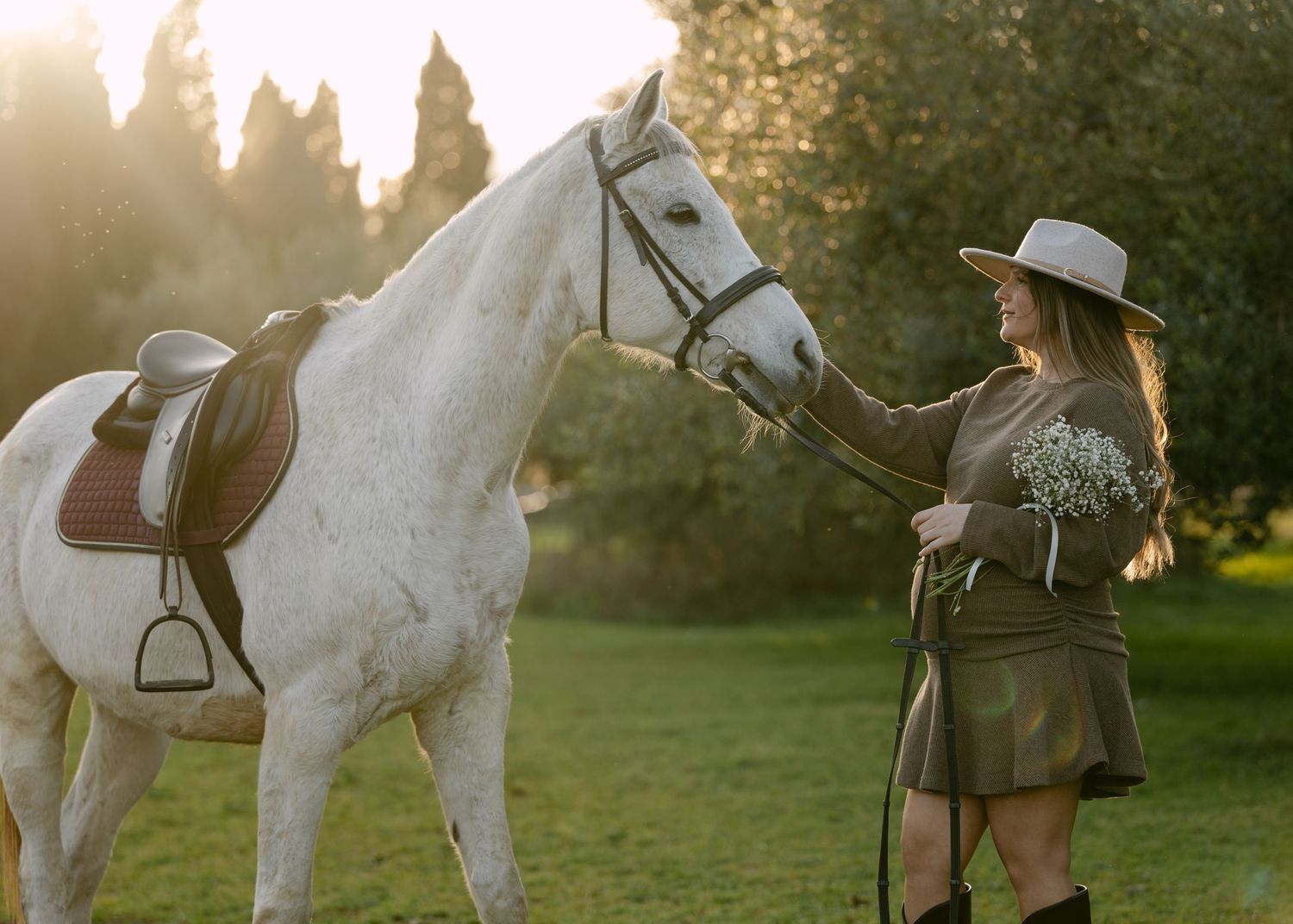 Woman in hat and riding boots holding bouquet while leading white horse in sunny garden.