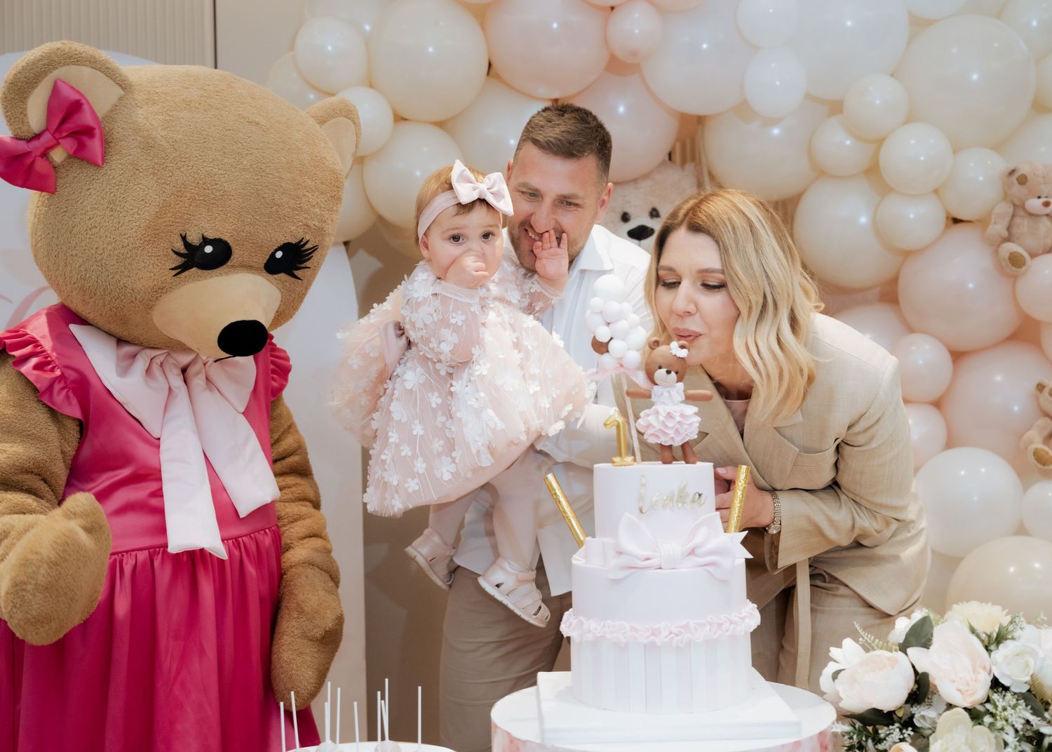 Family with baby girl blowing out candles on pink birthday cake beside a person in bear costume.