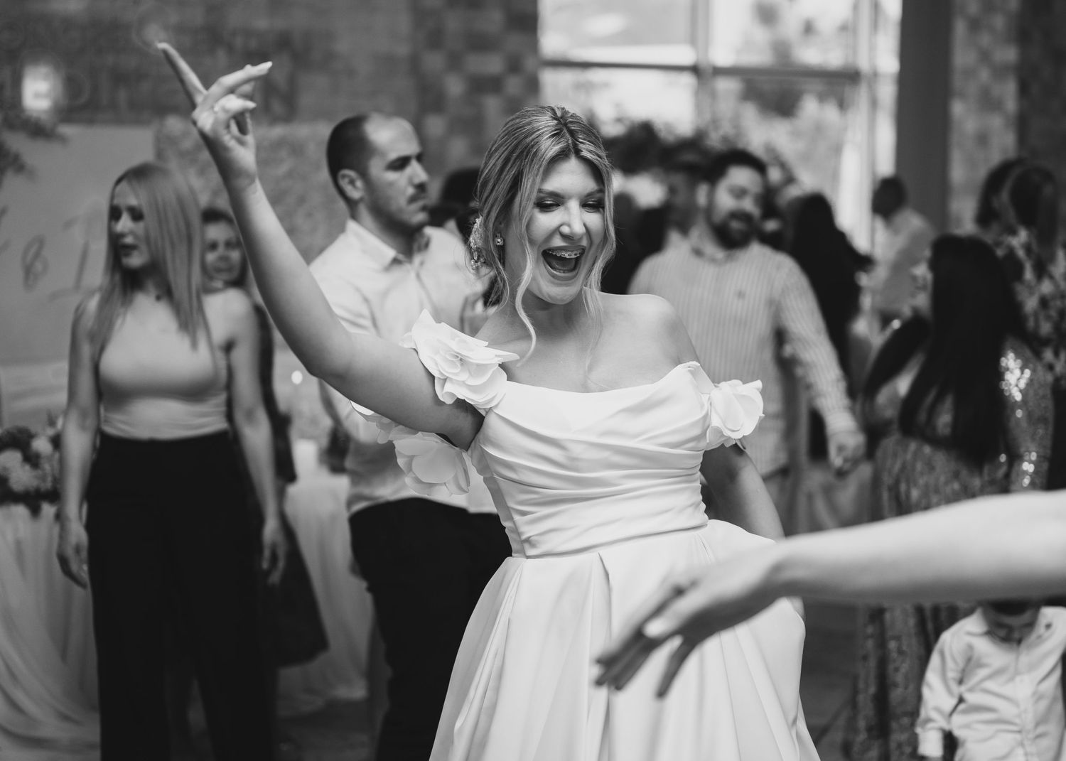 Joyful bride in off-shoulder white gown dancing energetically at wedding reception in black and white.