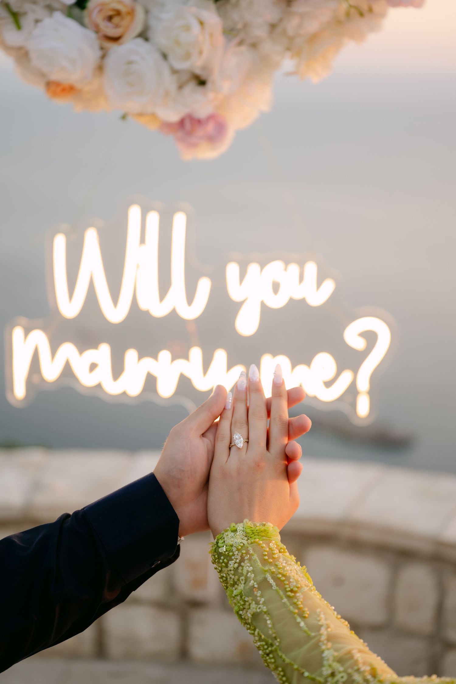 Couple's hands touching with engagement ring near neon Will You Marry Me sign and floral arrangement.
