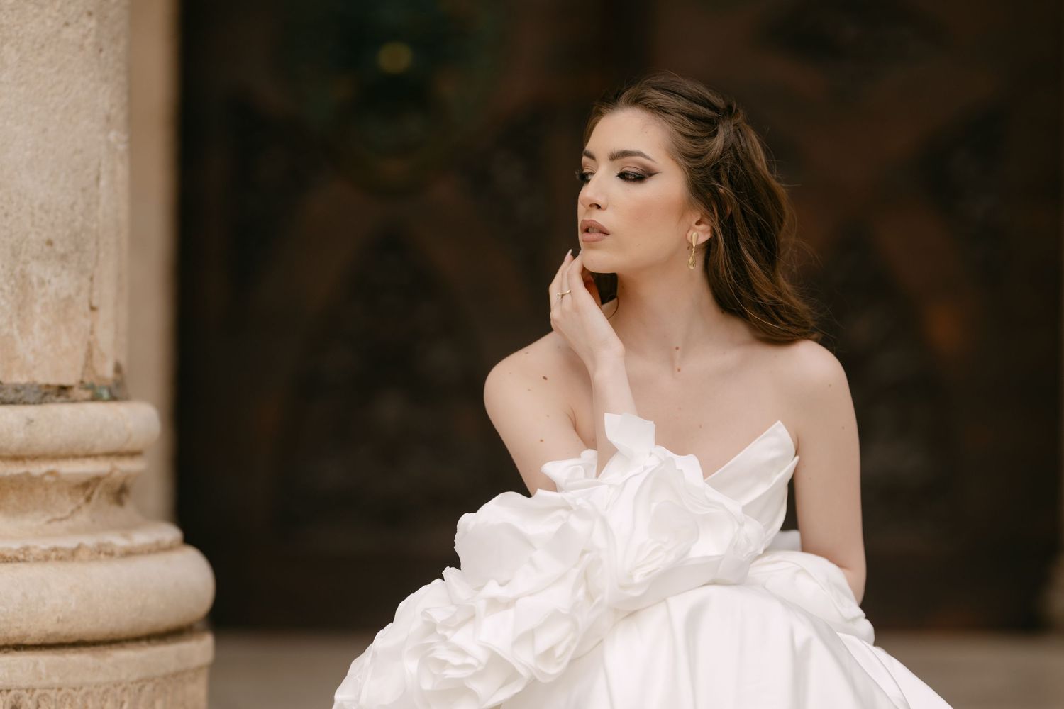 Bride in a strapless white ruffled gown sitting pensively beside a stone column in soft light.