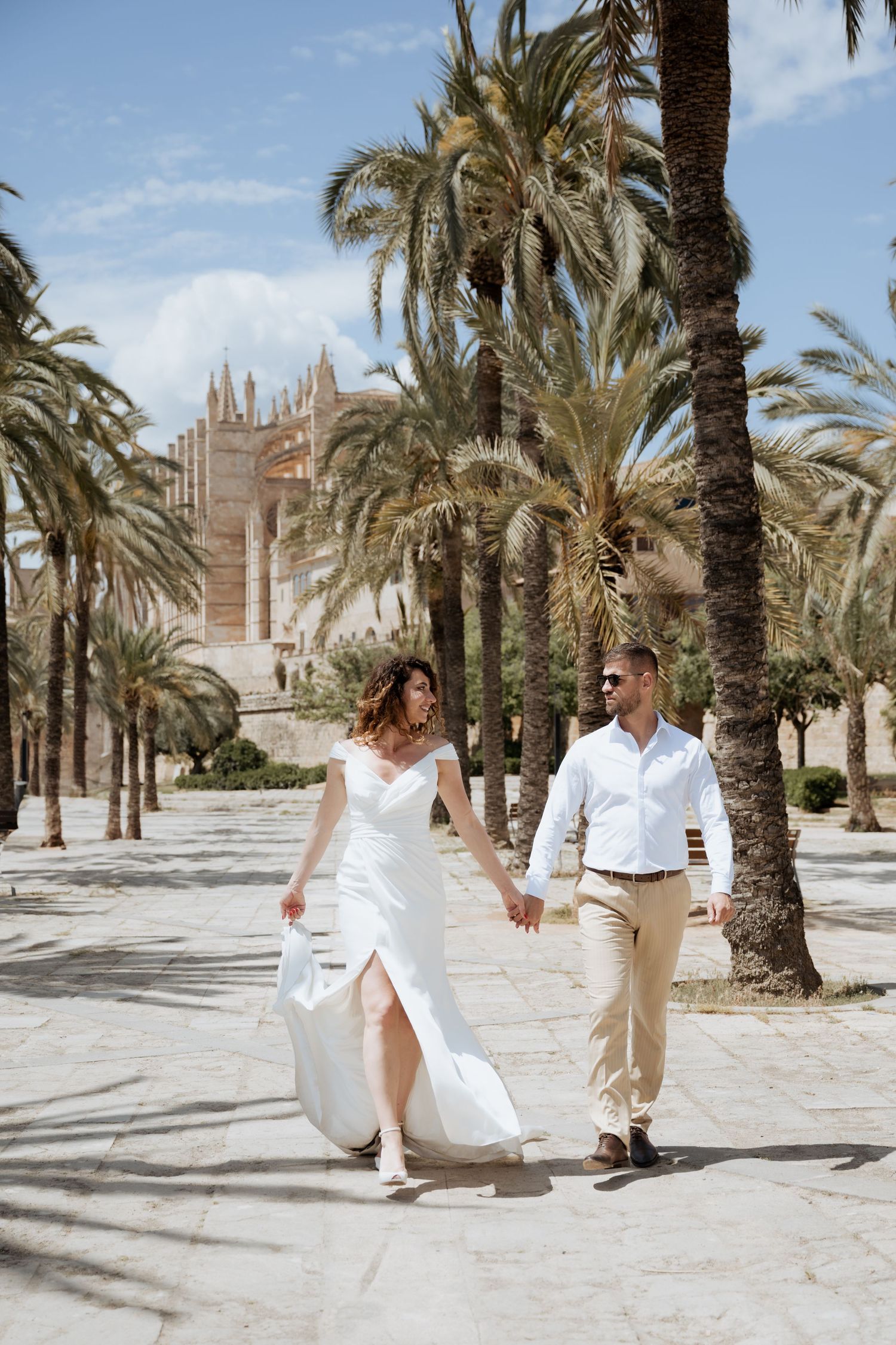Wedding couple walks hand-in-hand among tall palm trees with historic cathedral in background.