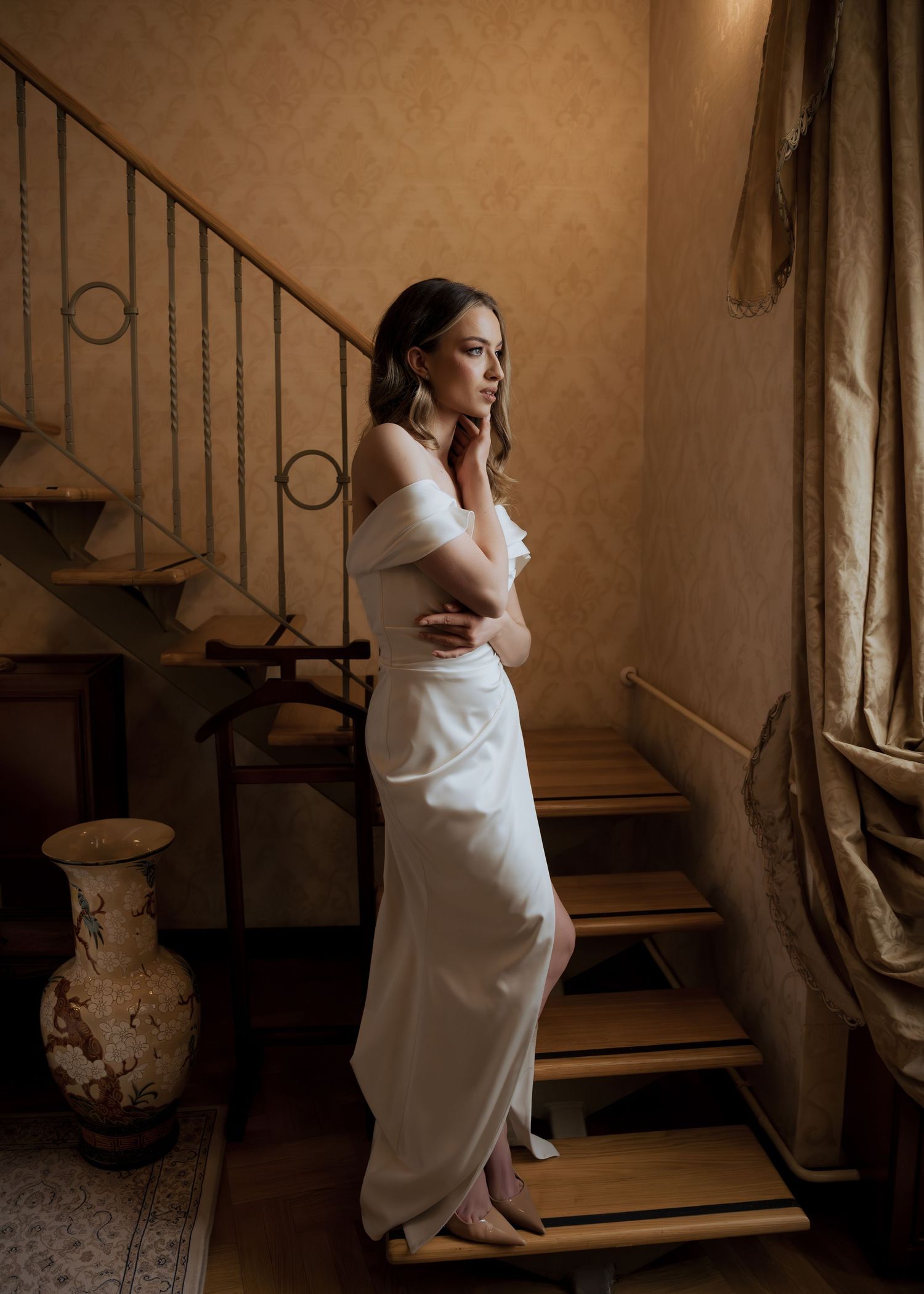 Elegant bride in off-shoulder white gown poses thoughtfully on wooden staircase in warm interior.