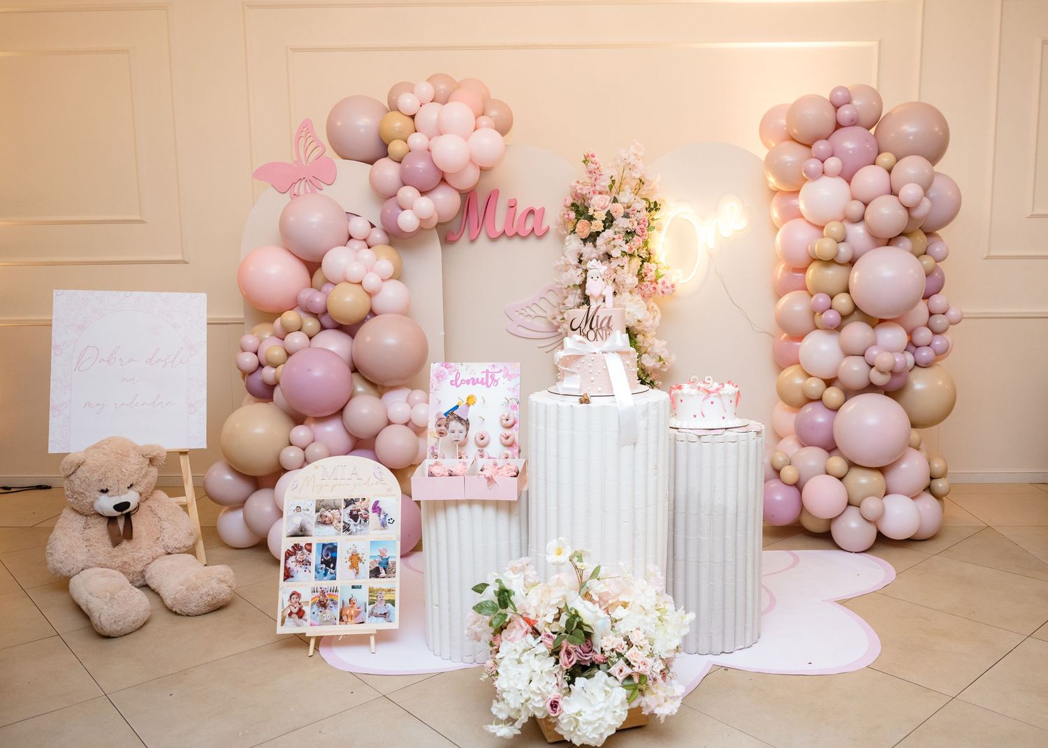 Pink and white first birthday party dessert table with balloon columns and floral decorations.