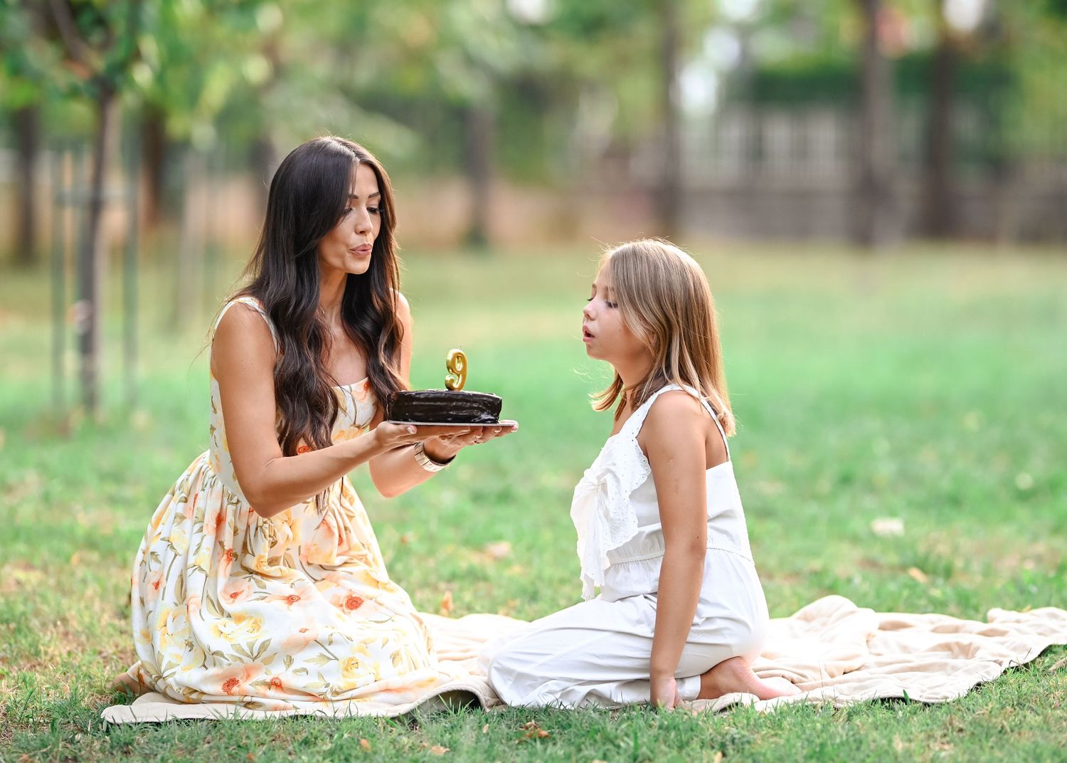 Excited young girl blowing out birthday candle as mother presents chocolate cake outdoors.