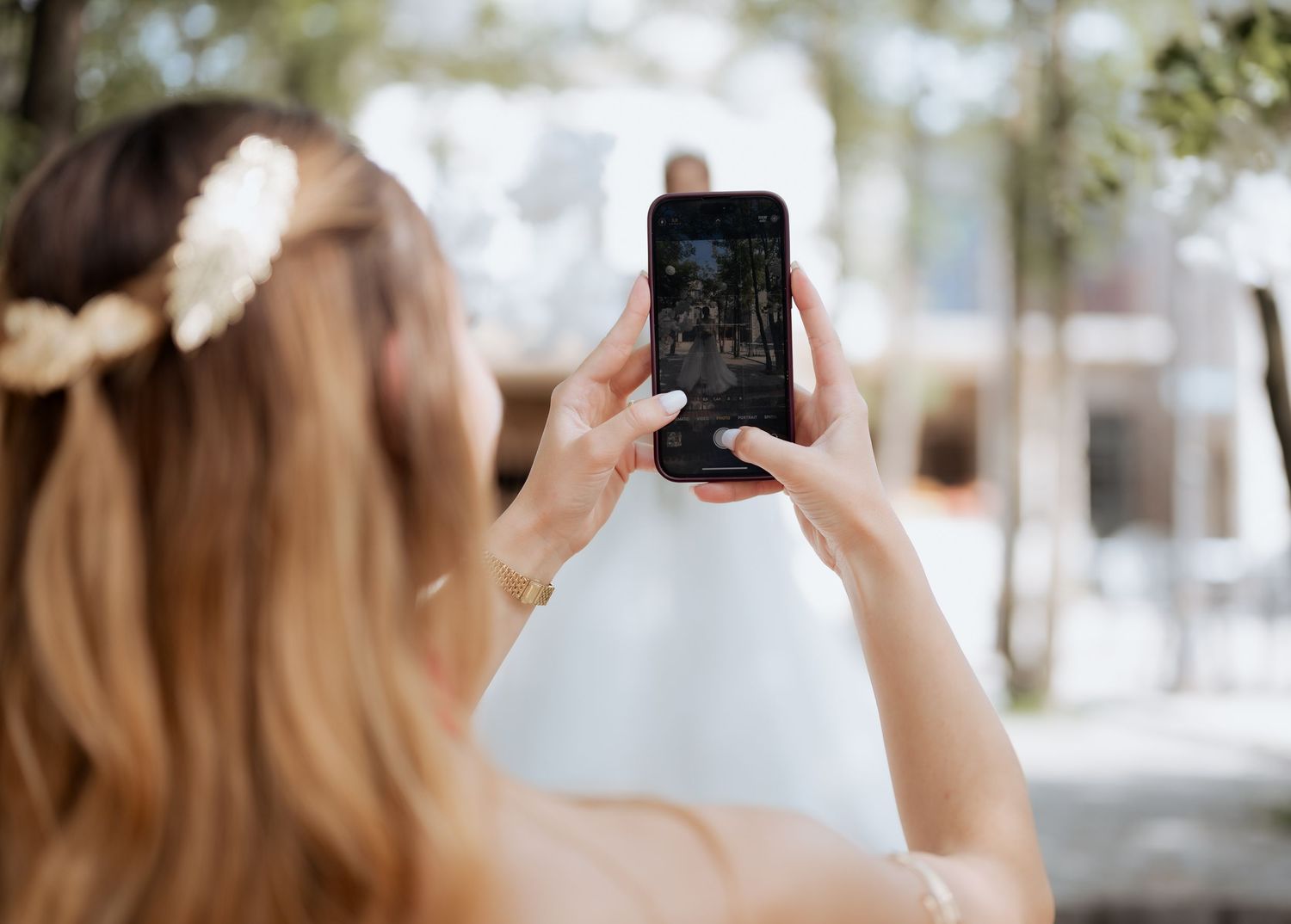 Wedding guest with floral hair accessory photographing ceremony on smartphone at outdoor wedding.