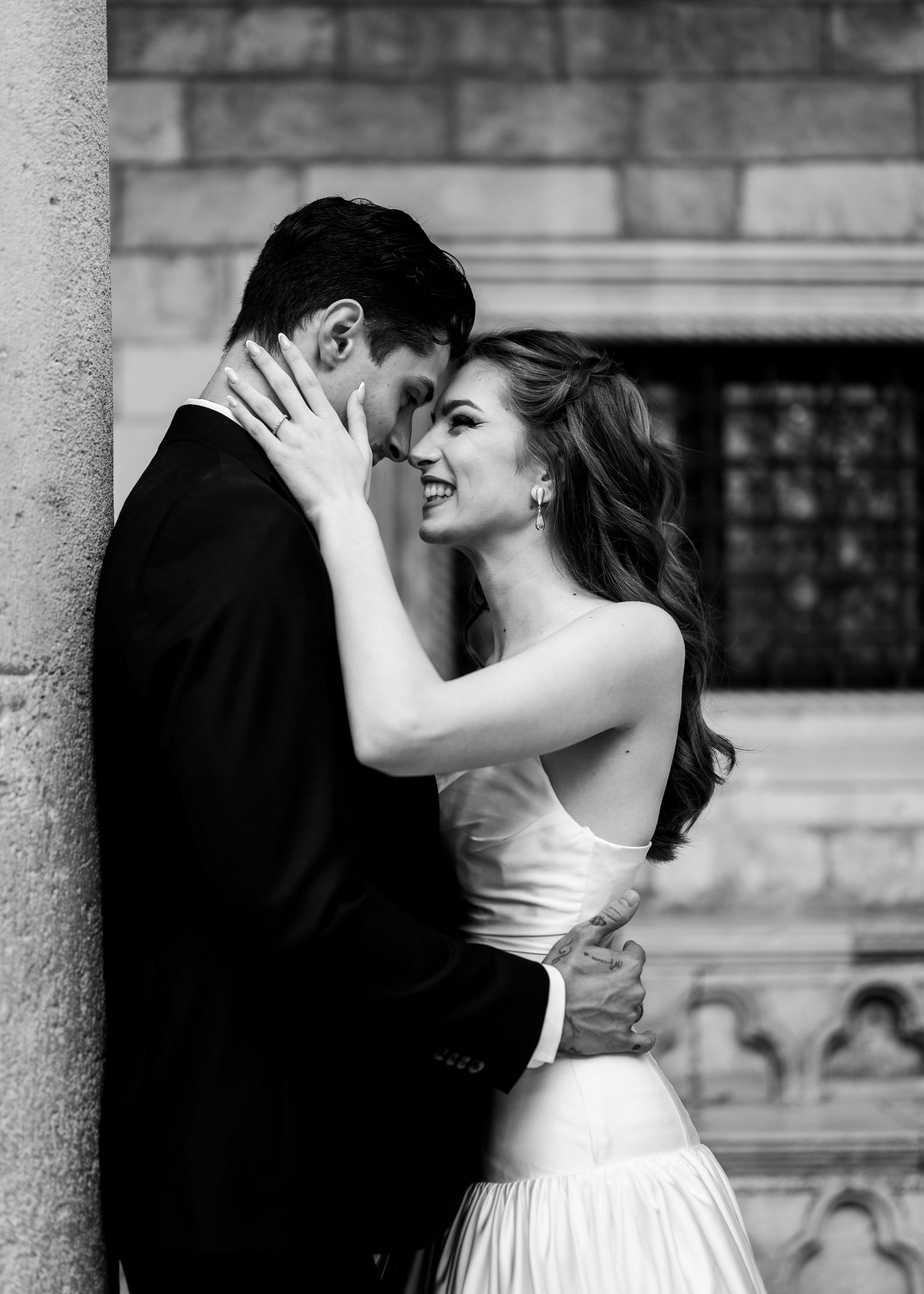 Romantic black and white photo of a smiling bride embracing her groom against a stone column.