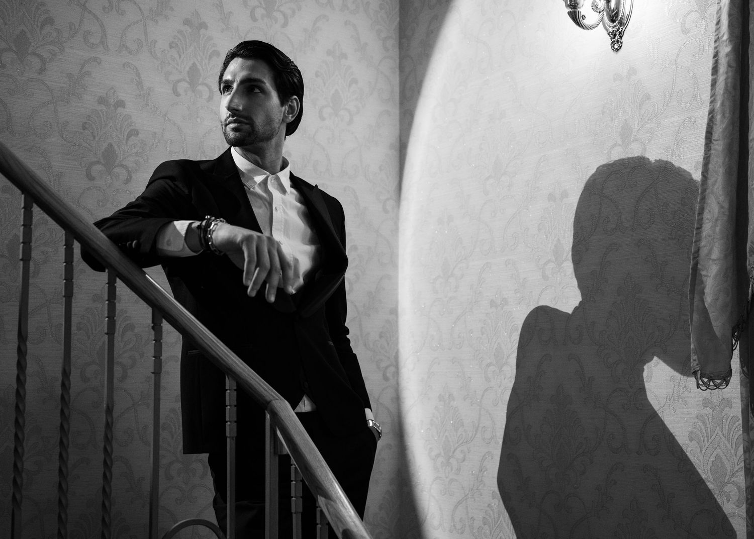 Black and white artistic photo of groom on staircase with bride's shadow on ornate wall.
