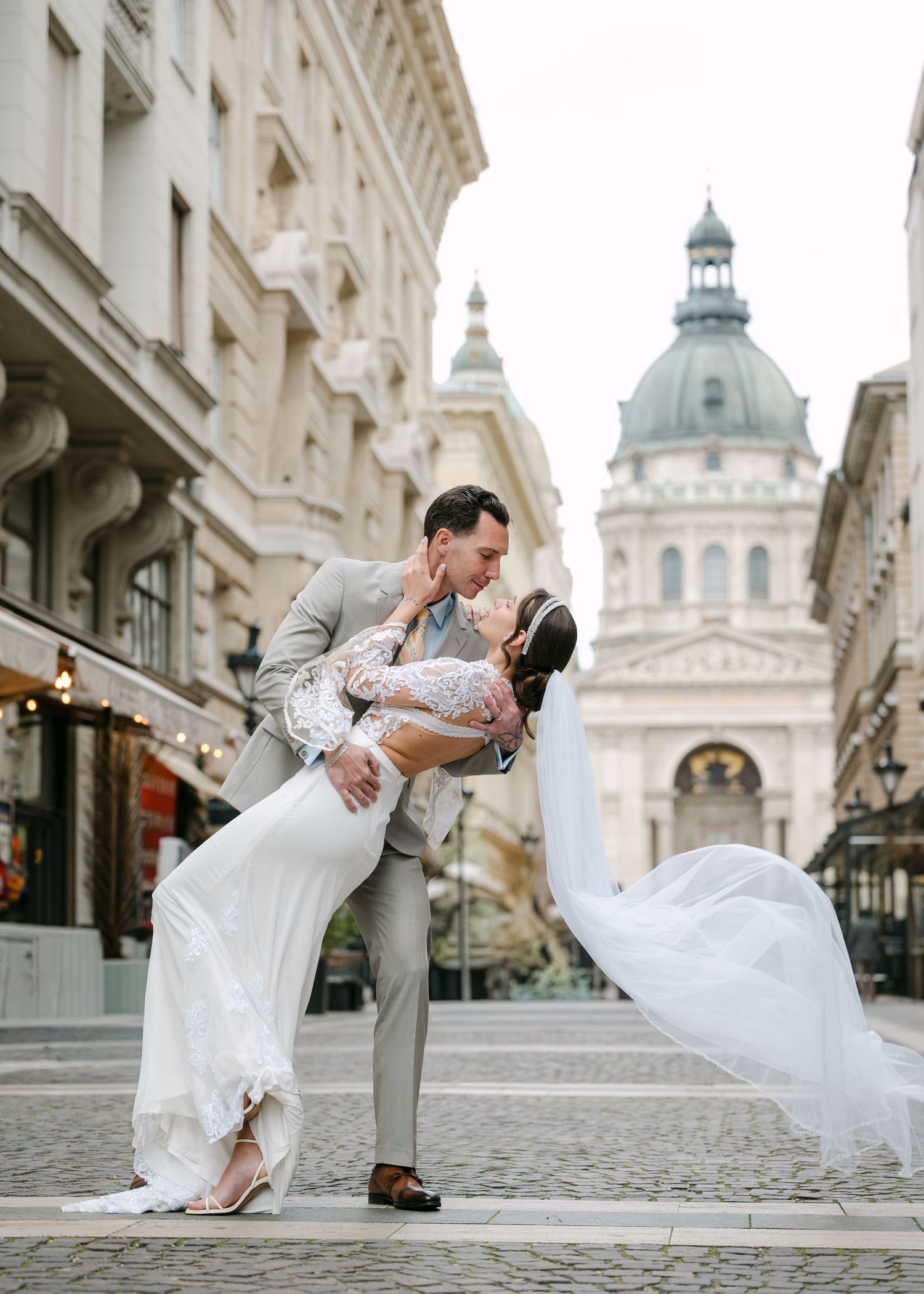 Groom dipping bride for romantic kiss on cobblestone street with Budapest basilica backdrop.