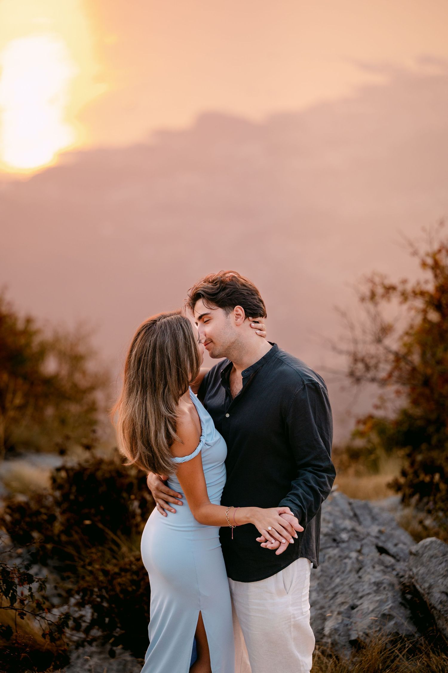 Couple sharing a romantic kiss at golden sunset with mountain silhouettes in warm glowing background.