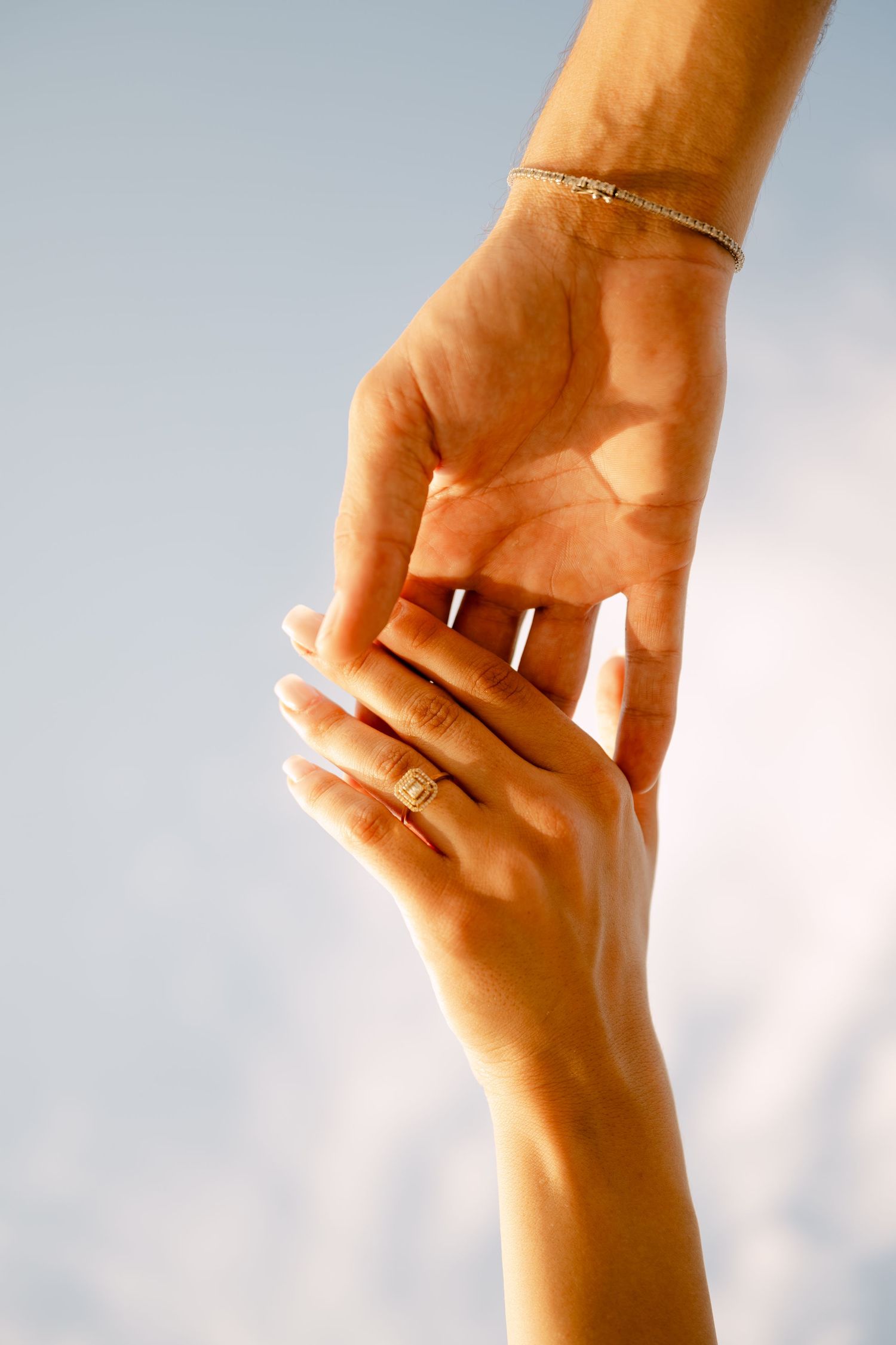 Close-up of two hands gently touching against soft blue sky with gold bracelet and ring.