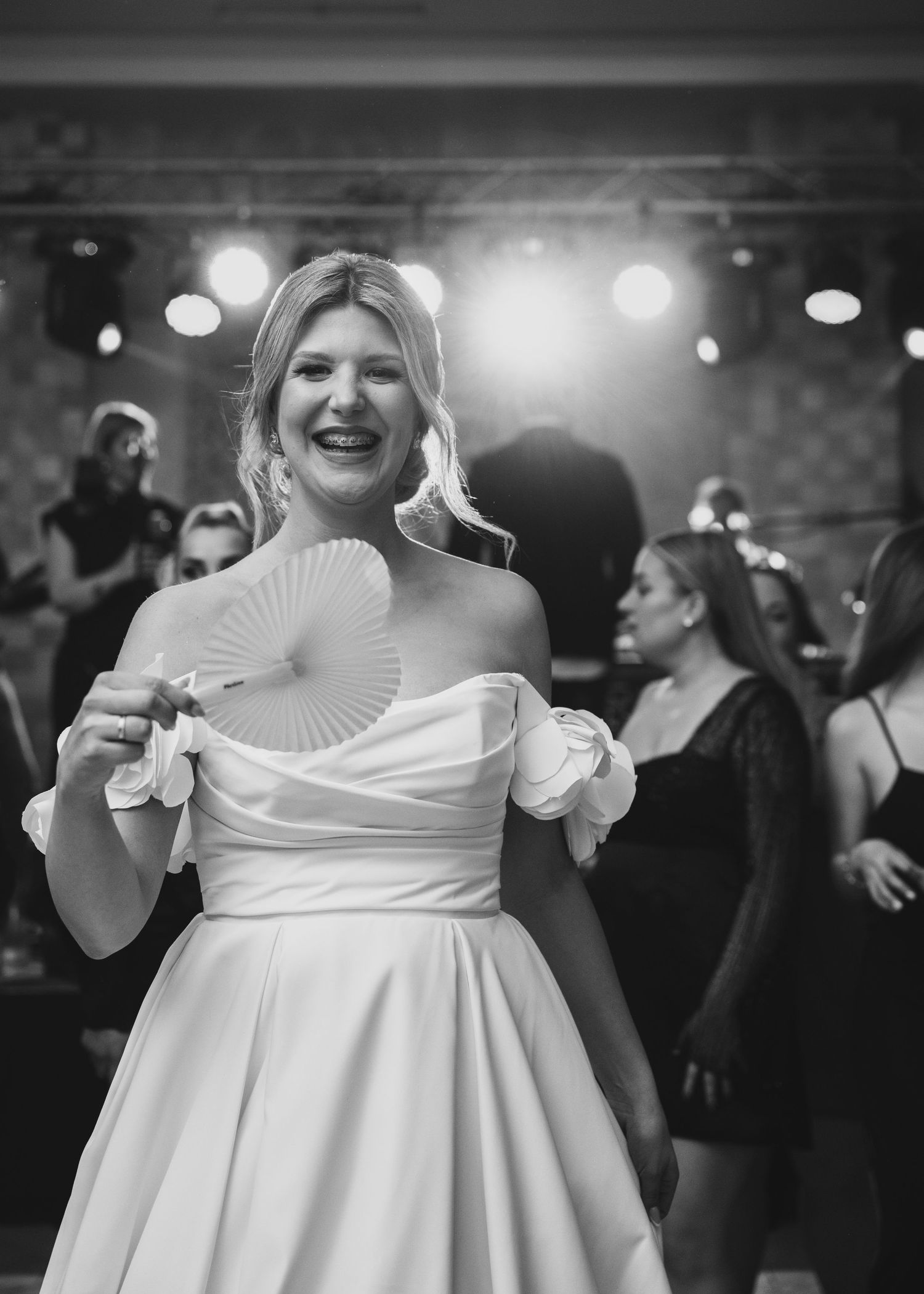 Smiling bride in white off-shoulder gown holding decorative fan at lively wedding reception.