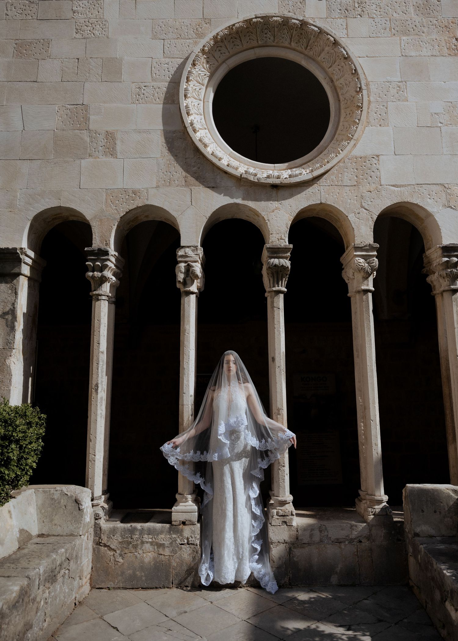 Bride in lace wedding gown with flowing veil stands before ornate Romanesque arches and rose window.