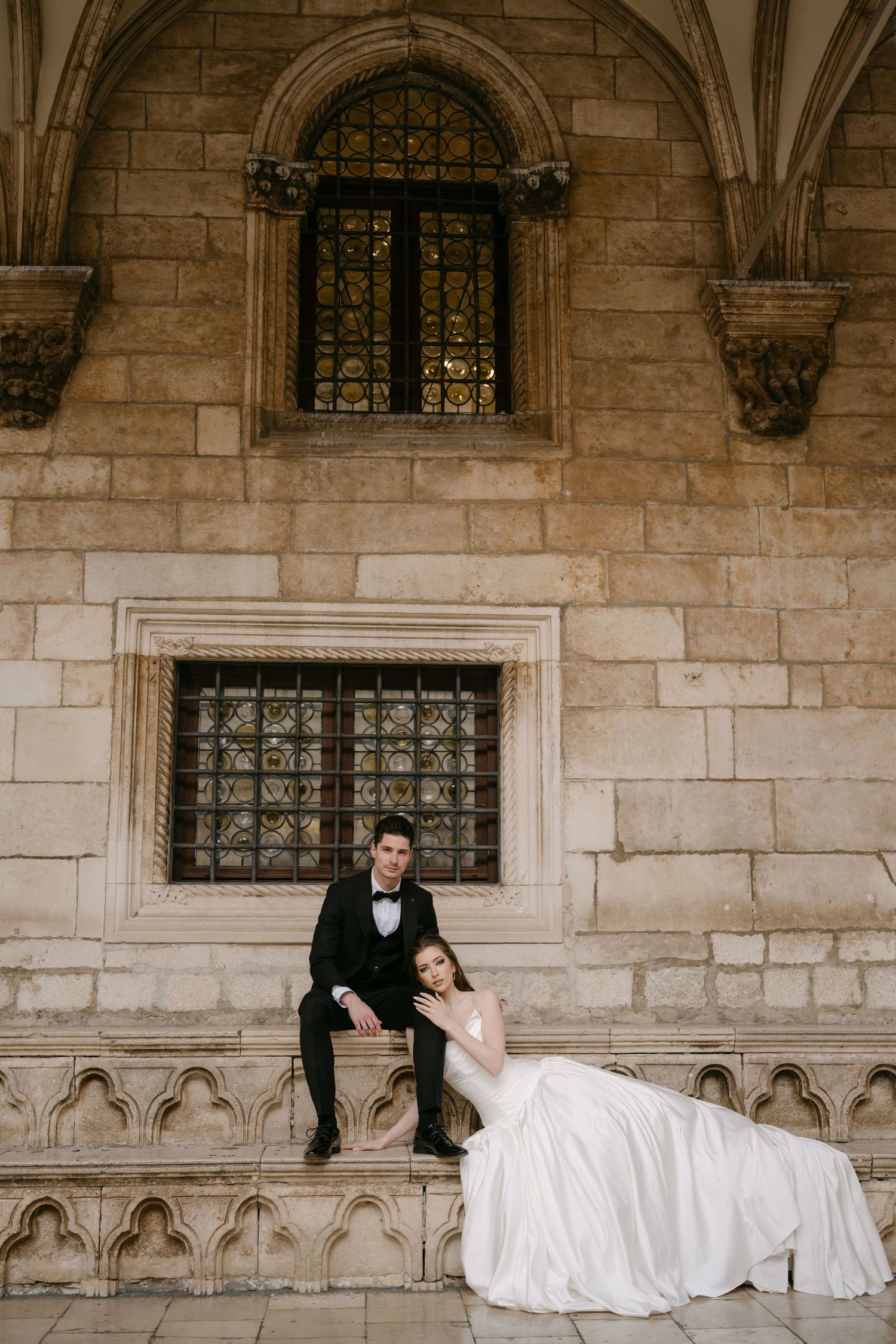Bride and groom seated on ornate stone steps beneath a Gothic window of a historic building.