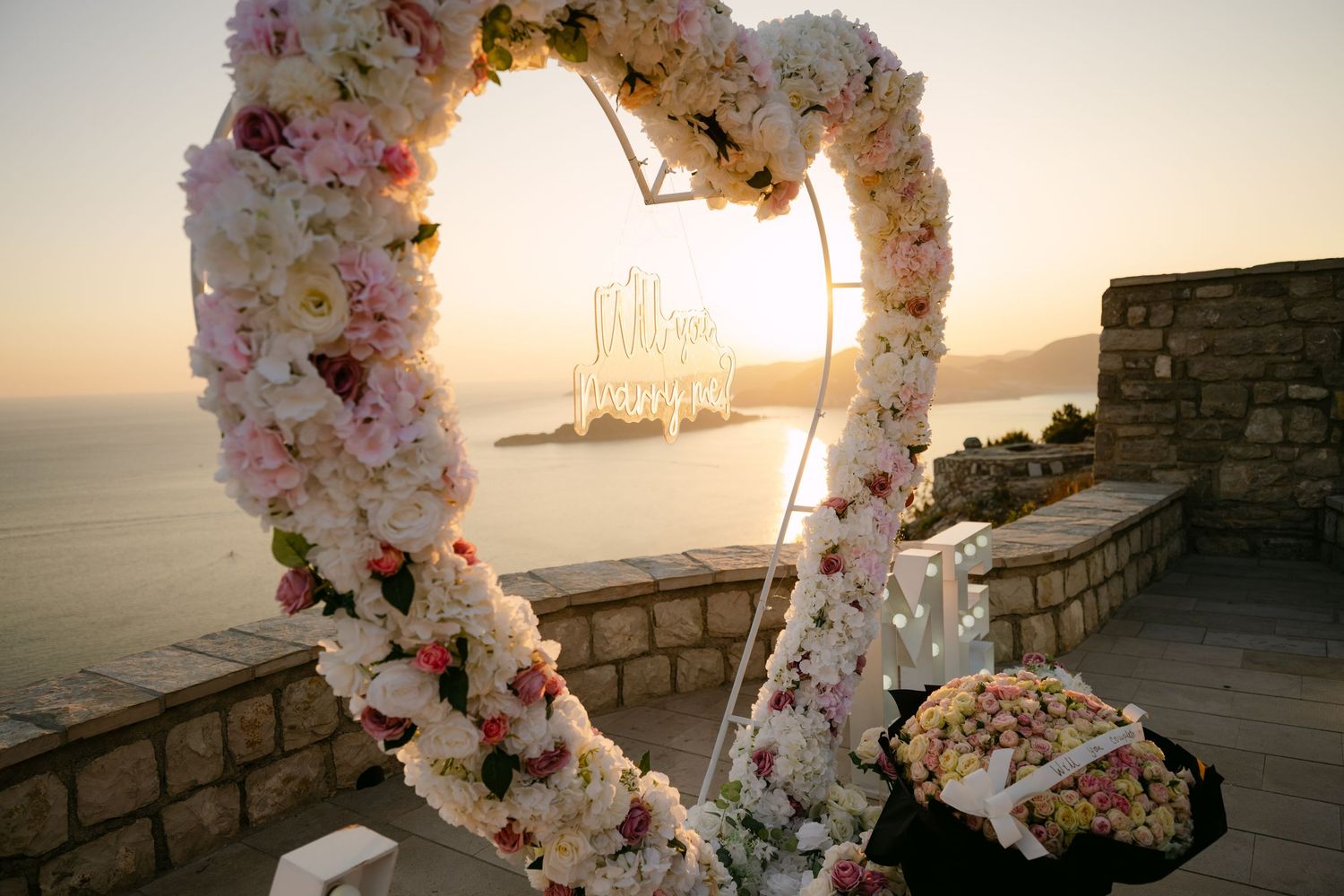 Beautiful floral heart arch proposal decoration glowing at sunset with ocean and chandelier backdrop.