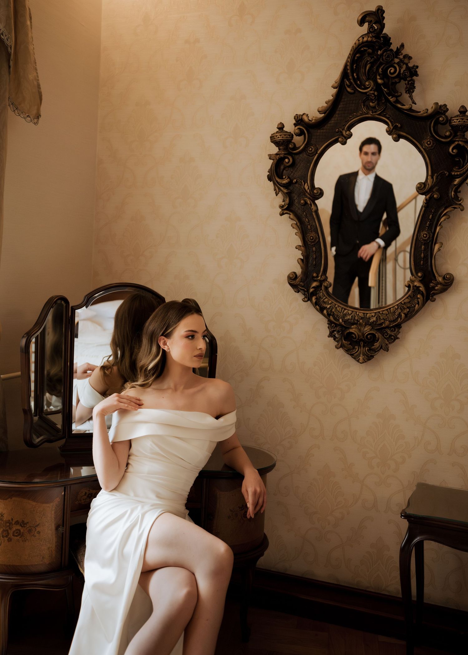 Bride in off-shoulder white gown sits elegantly while groom's reflection appears in an ornate gold-framed mirror.
