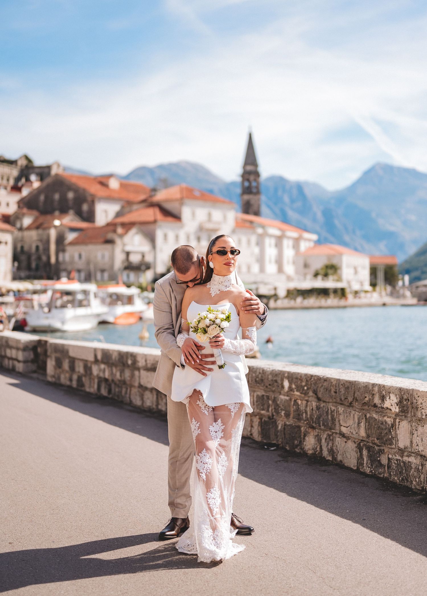 Bride in lace wedding dress holding bouquet with groom embracing her by Montenegro waterfront.