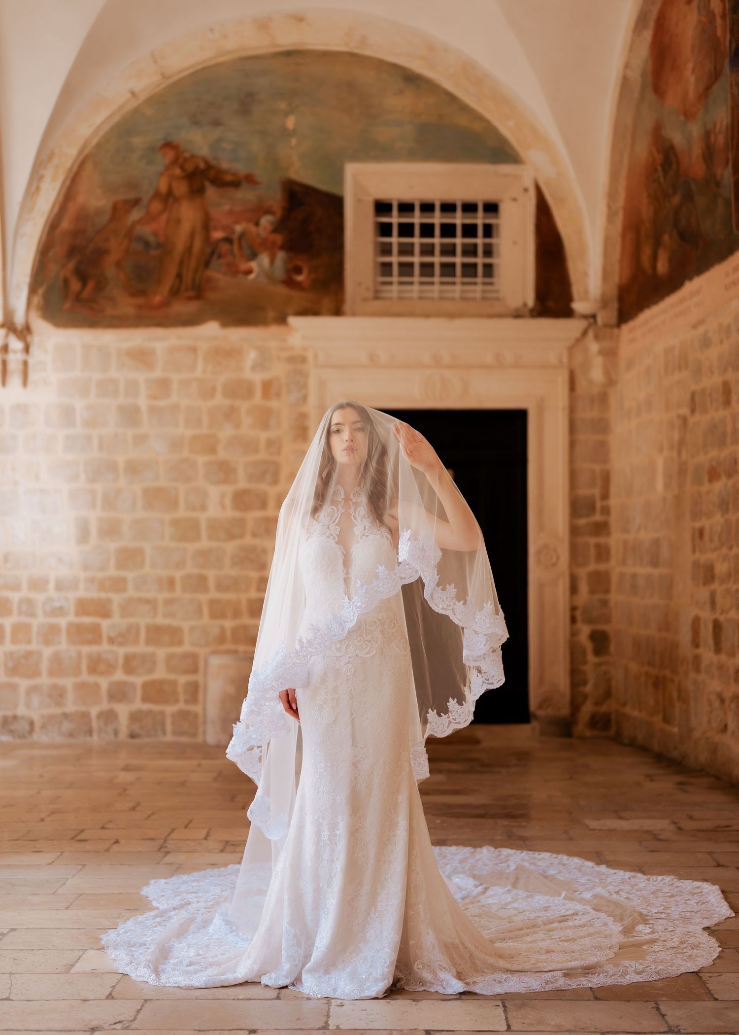 Elegant bride in lace gown and long veil stands beneath arched stone ceiling with Renaissance fresco paintings.