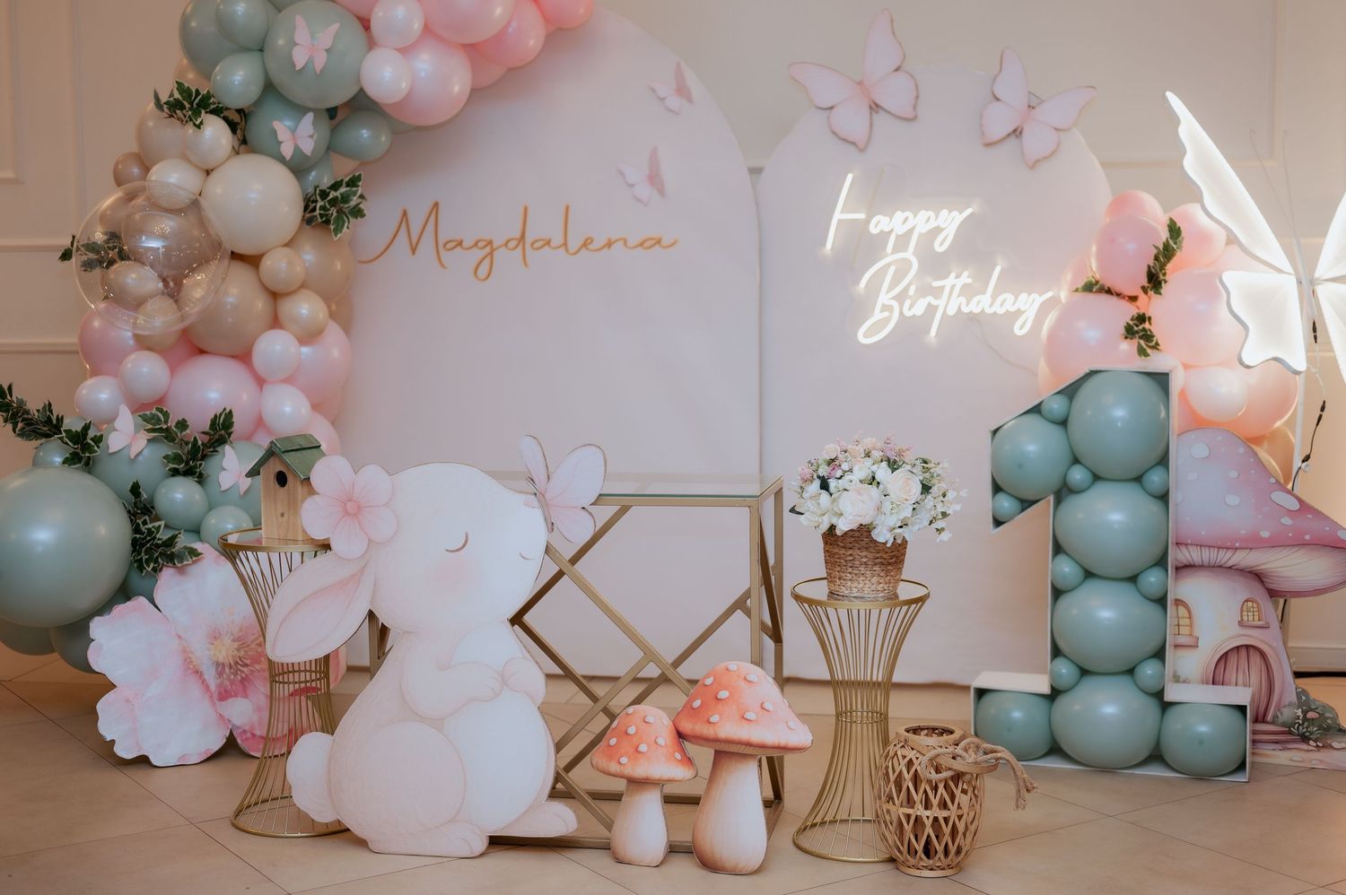 Pastel butterfly-themed first birthday party setup with sage balloons, mushroom props, and glowing Happy Birthday neon sign.