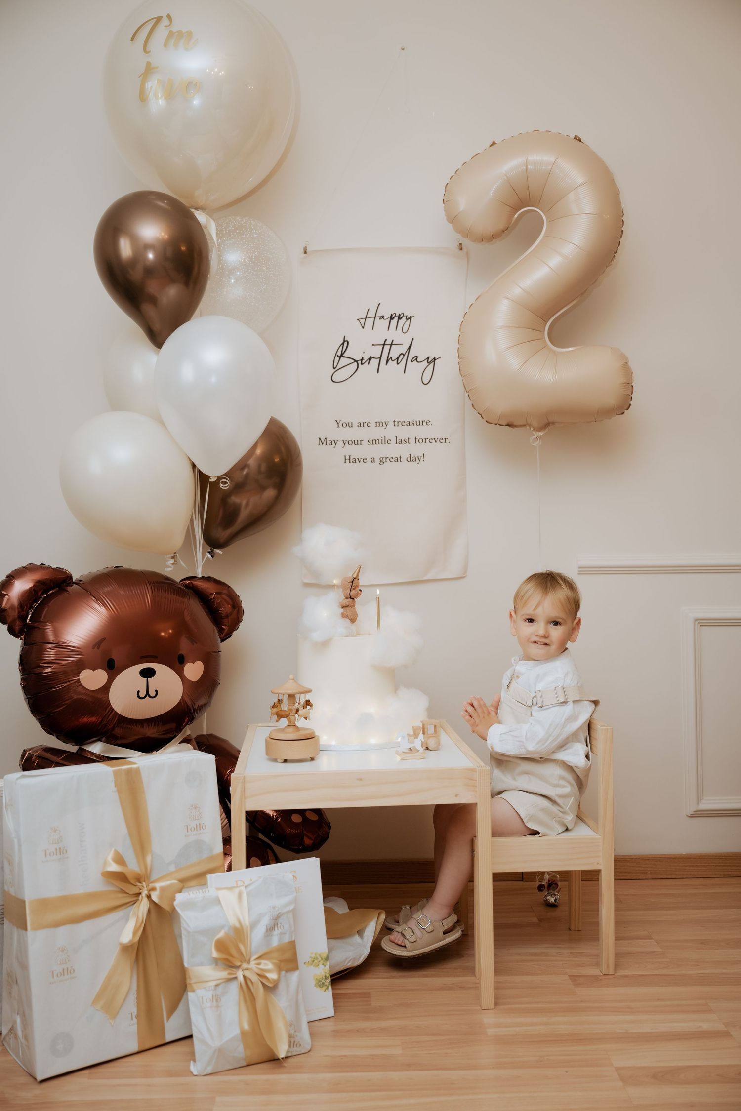 Toddler celebrates 2nd birthday with white cake, bear balloon, gifts, and brown and white balloon decorations.
