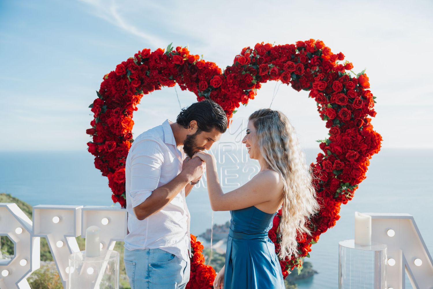 Couple holding hands and smiling in front of a large red rose heart arch with 'Marry Me' marquee letters.